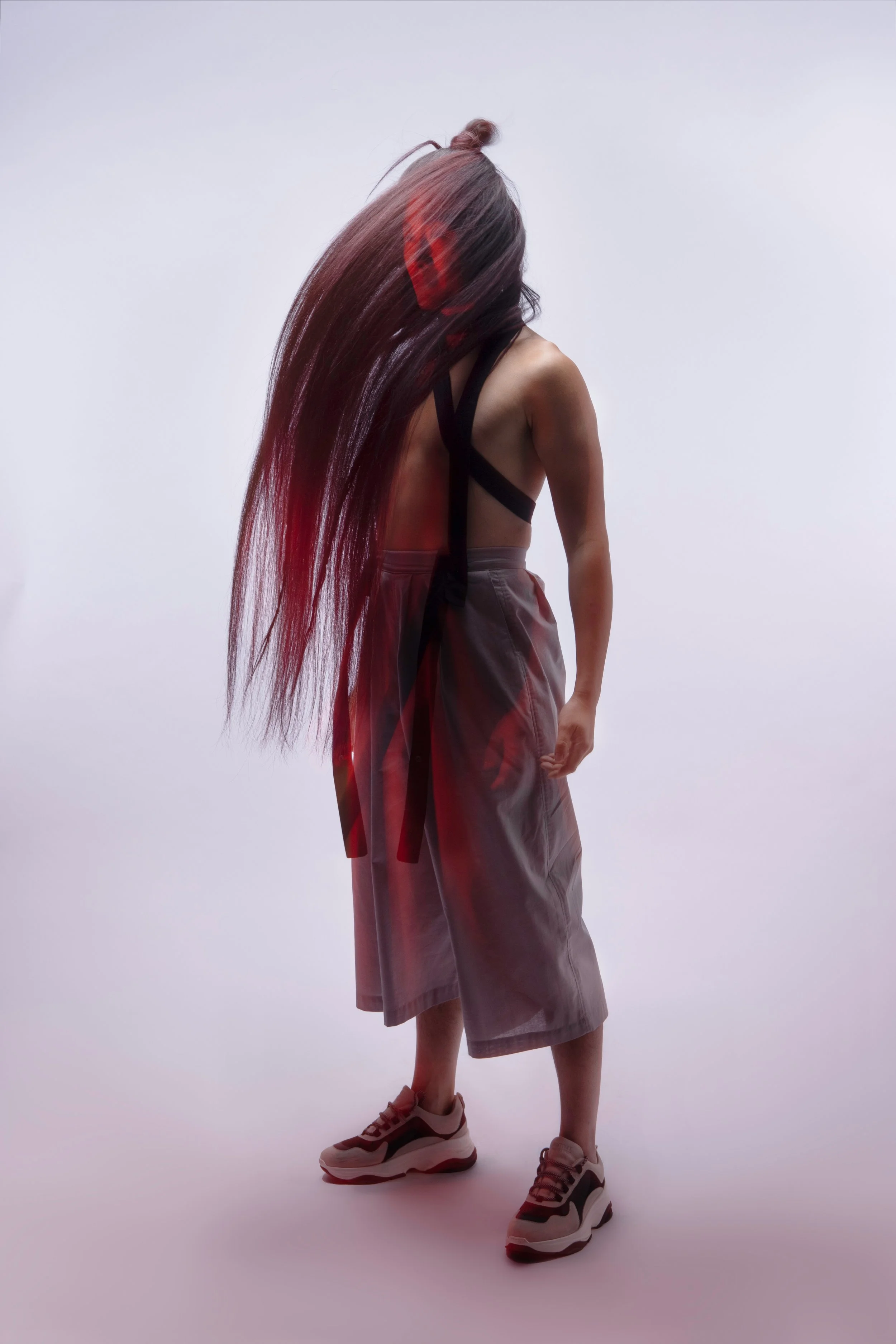 A woman with long dark hair and light skin, standing against a plain white background, wearing a black strappy top, gray wide-legged pants, and sneakers, with her head bent forward.