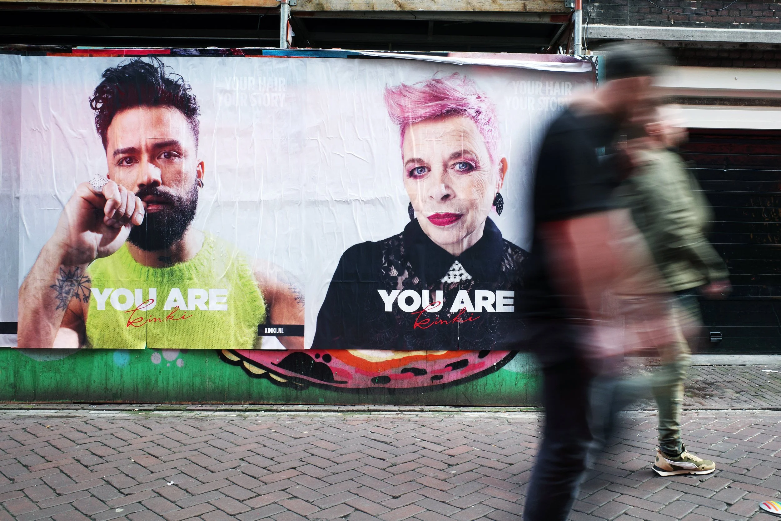 Street scene with blurred pedestrians walking past a large billboard featuring two portraits and text. The billboard displays a man with dark hair and a beard wearing a bright green tank top, and a woman with short pink hair and bold makeup, dressed 