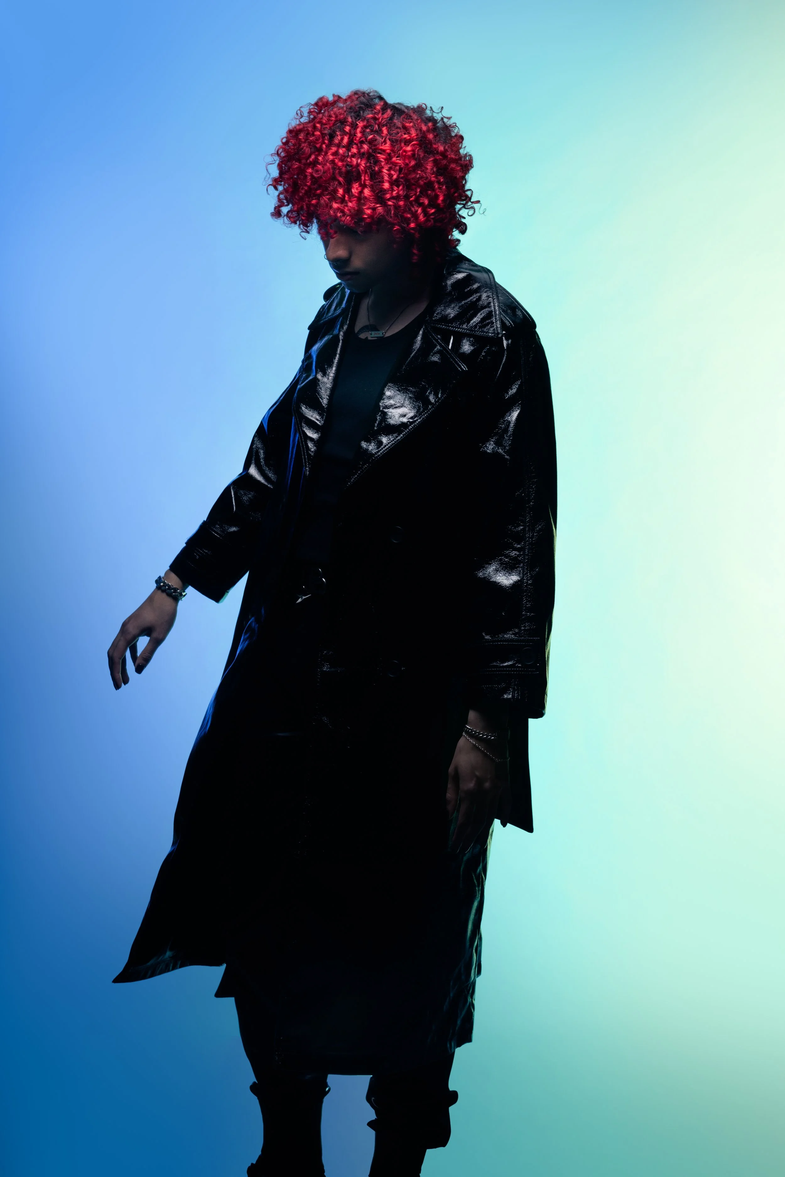 Person with curly red hair wearing a shiny black leather coat, standing against a gradient blue and green background.