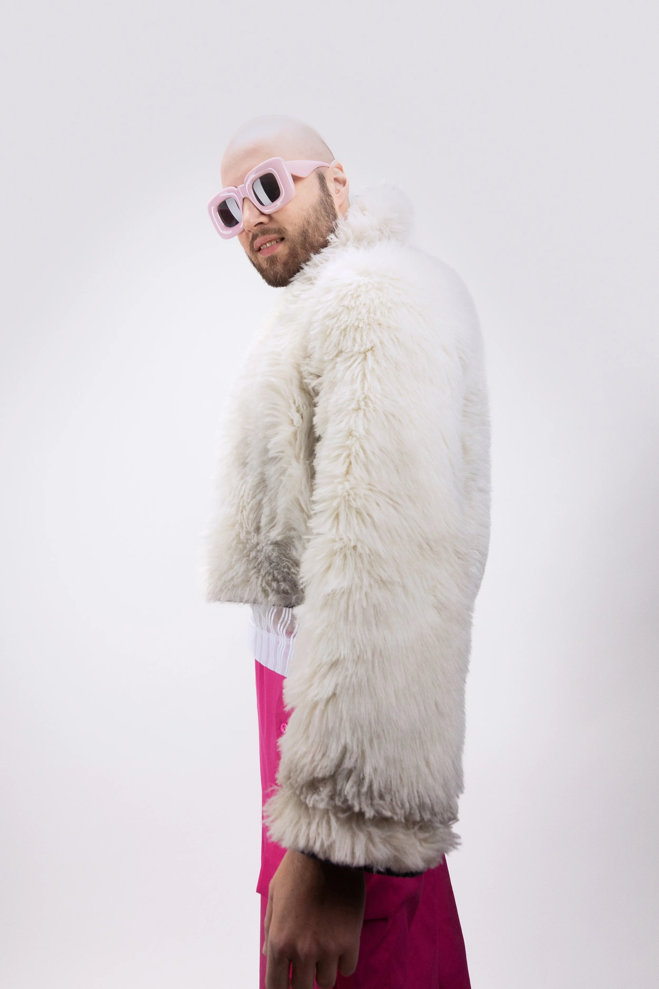 A man with a bald head and beard wearing pink sunglasses, a white faux fur coat, and pink pants, standing against a plain white background.