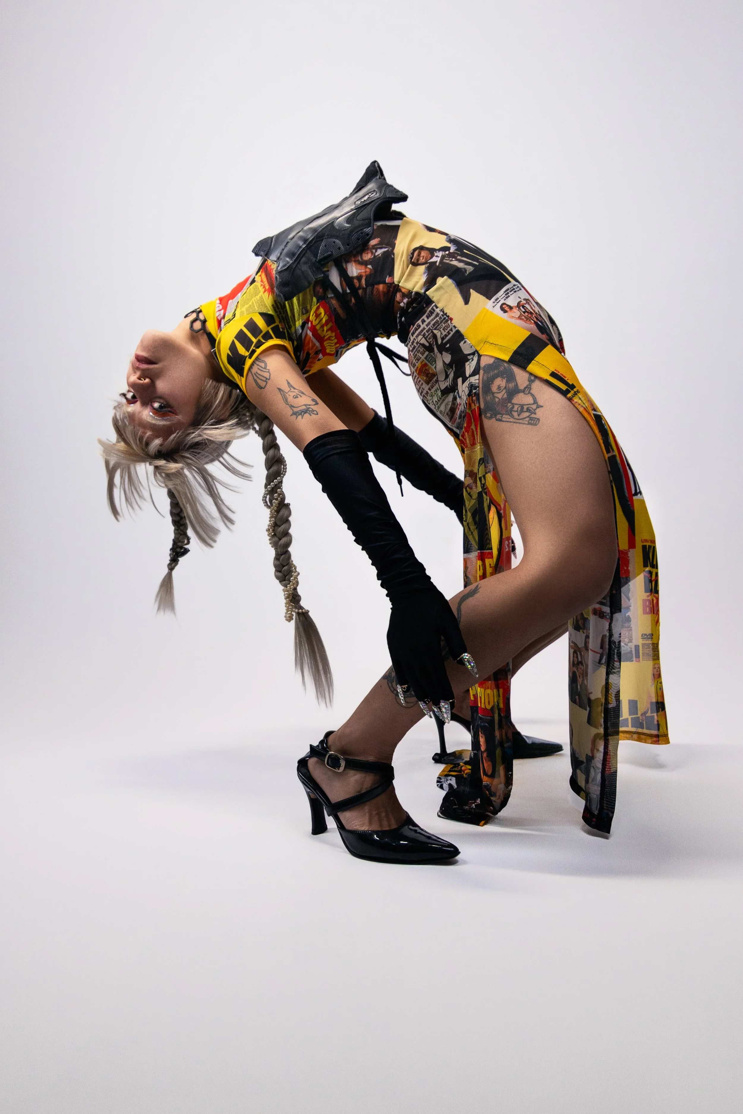 A woman with long braided blonde hair with beads, tattoos, and makeup, wearing a colorful comic book print dress, black gloves, and black high heels, posed in a backward bend against a plain white background.