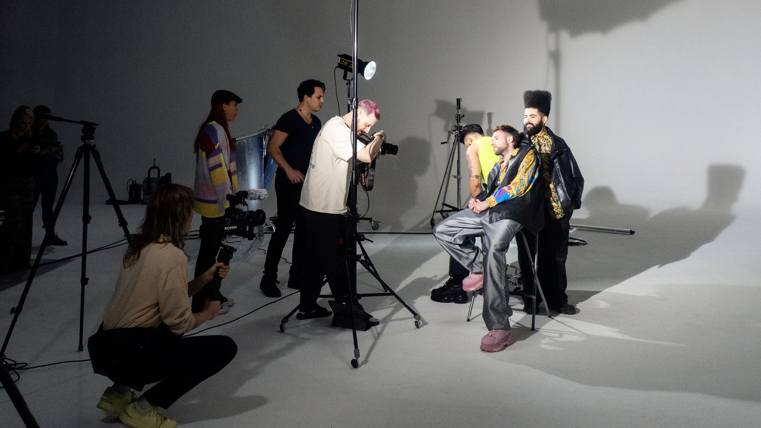 A photoshoot in a studio with three men and three women, some holding cameras and equipment, with two models sitting on stools and three people standing behind them. The models are dressed in colorful outfits, and there are professional lighting and tripods in the background.