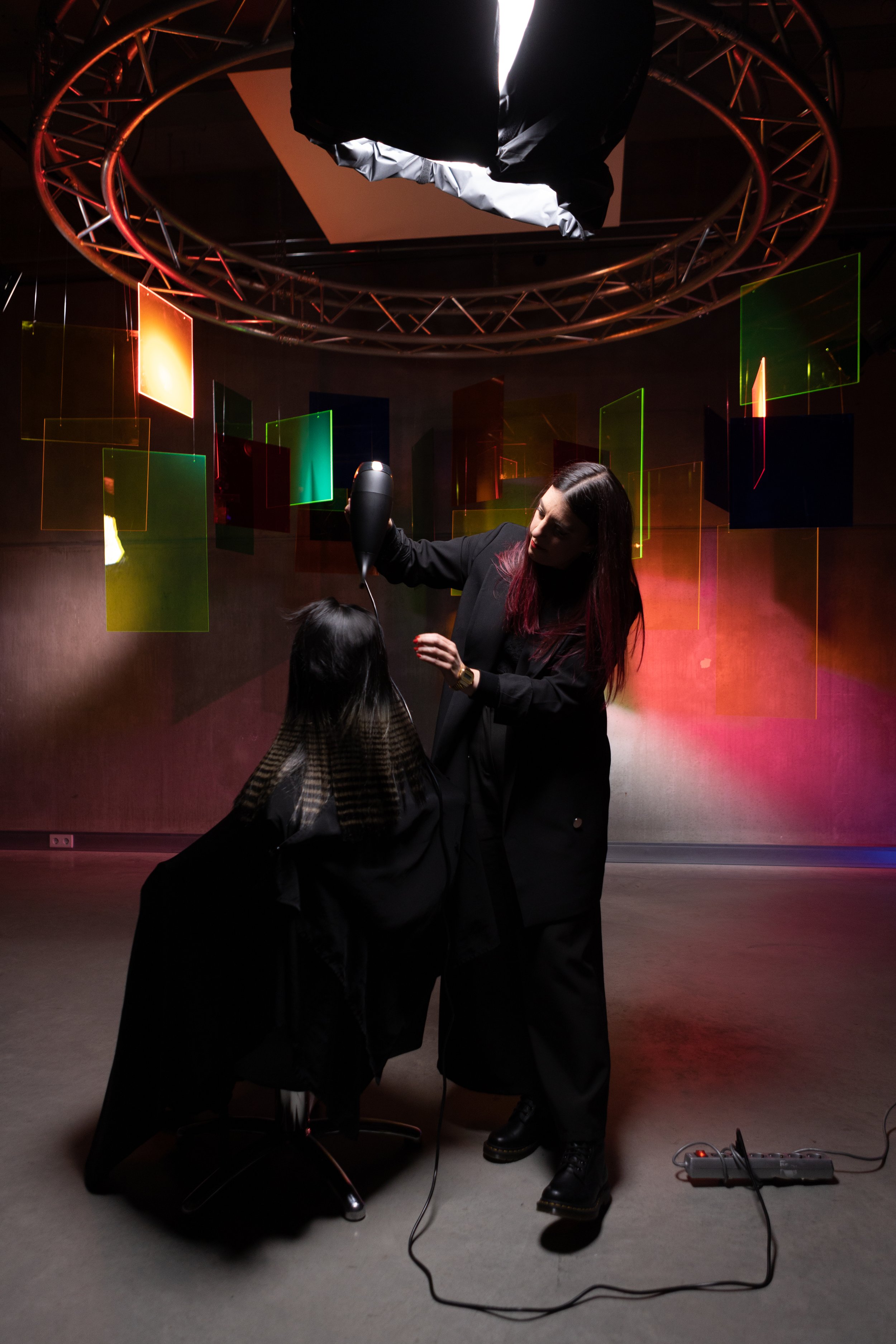 A woman is blow-drying a person's hair in a salon or studio with colorful, geometric light art on the background wall.