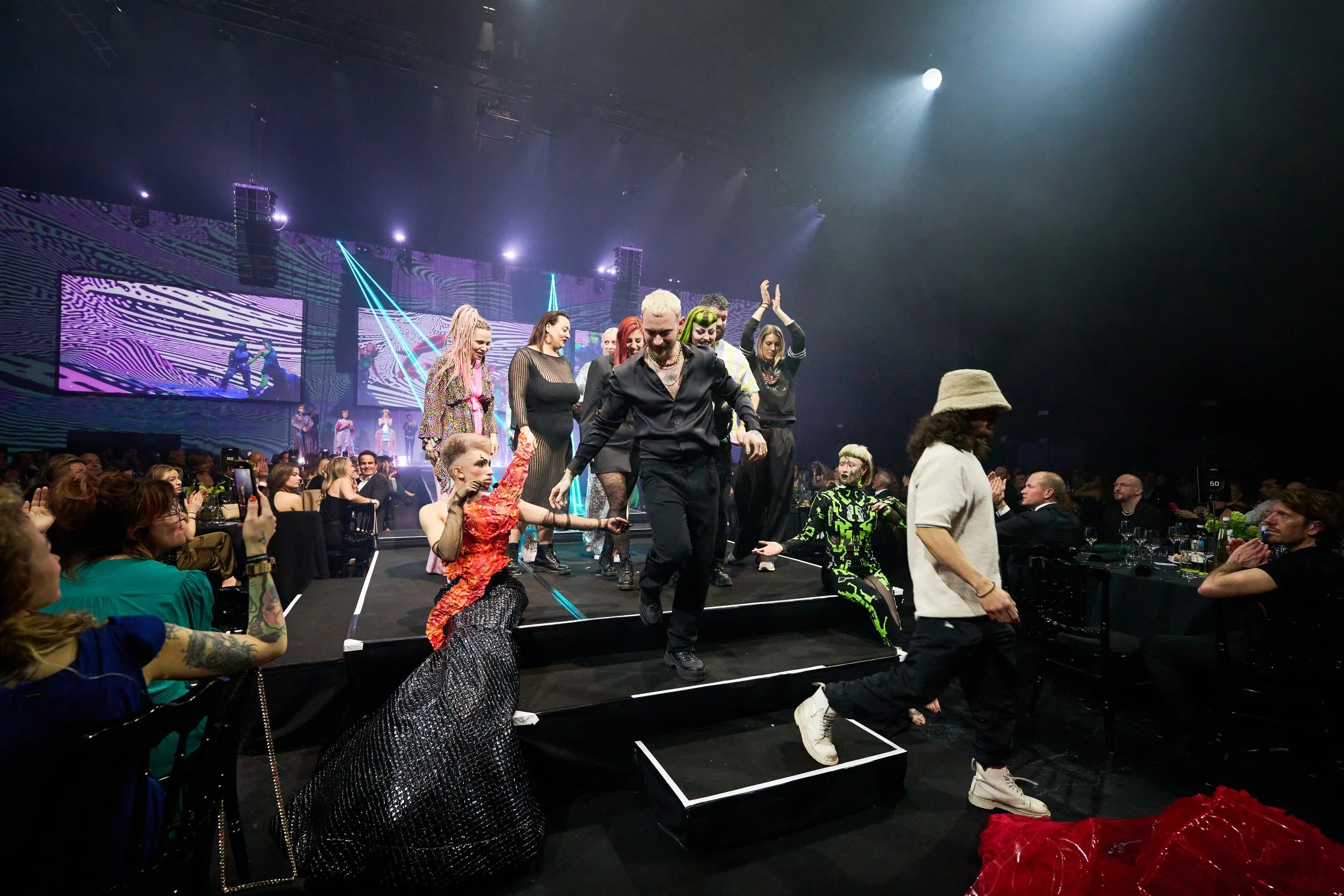People walking down a runway at a fashion show with a stage and colorful digital screens in the background, and audience seated on both sides.
