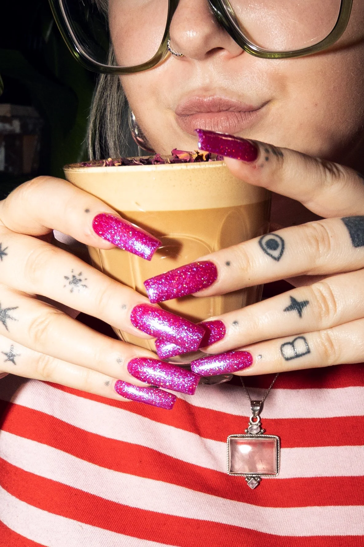 A woman with brightly colored, glittery pink nails, tattoos on her fingers, and a nose piercing is holding a coffee cup close to her face, wearing glasses, a red and white striped shirt, and a necklace with a large pendant.