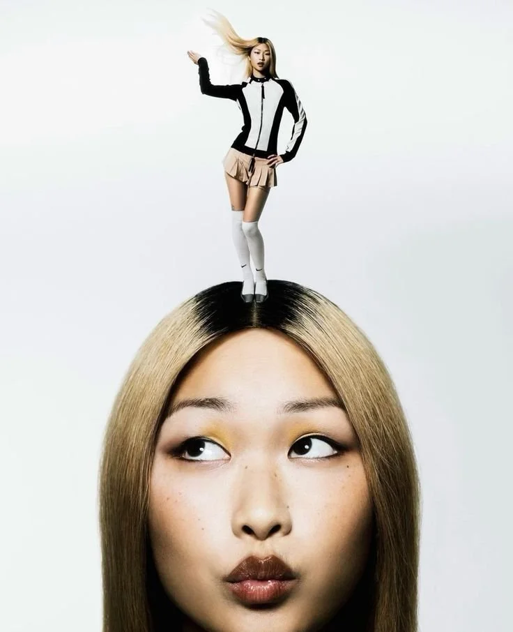 A large head of a woman with long blonde hair, looking sideways with a curious expression, and a tiny woman standing on top of her head.