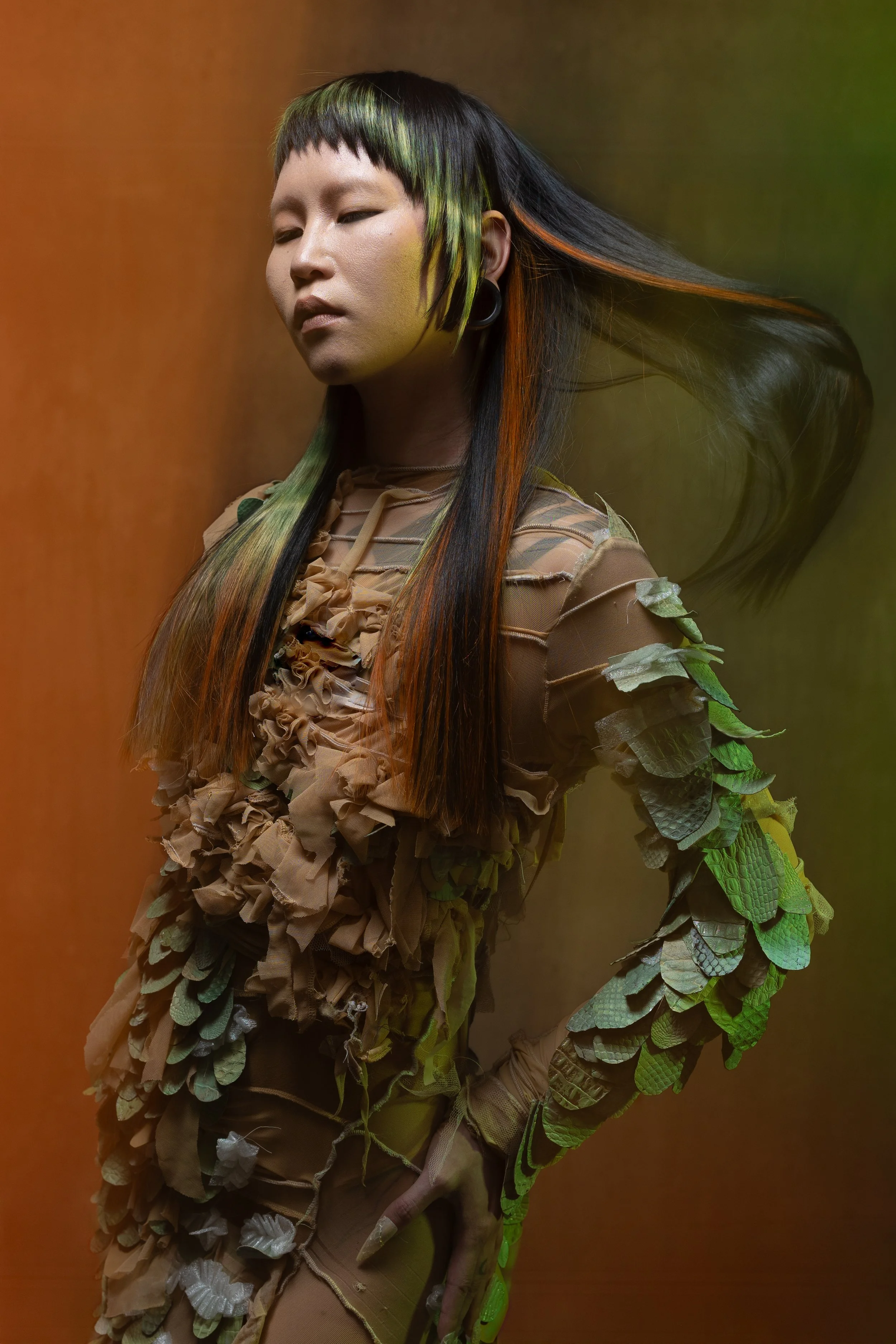 Person wearing a nature-inspired outfit with leafy details, posing with eyes closed against a blurred background.