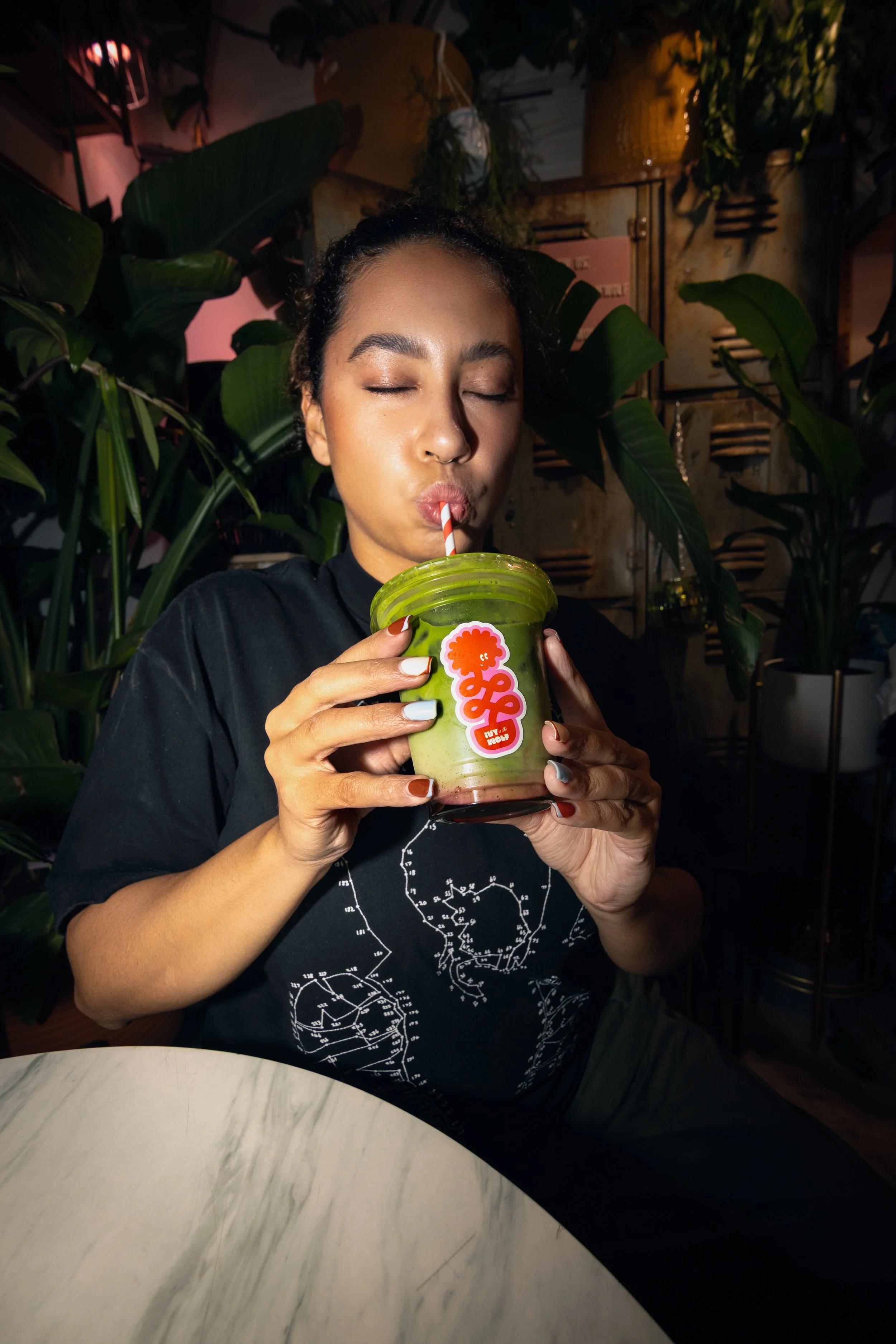 A woman with closed eyes drinks from a clear plastic cup with a green lid using a striped straw, holding the cup with both hands, in a setting with lush green plants and lockers in the background.