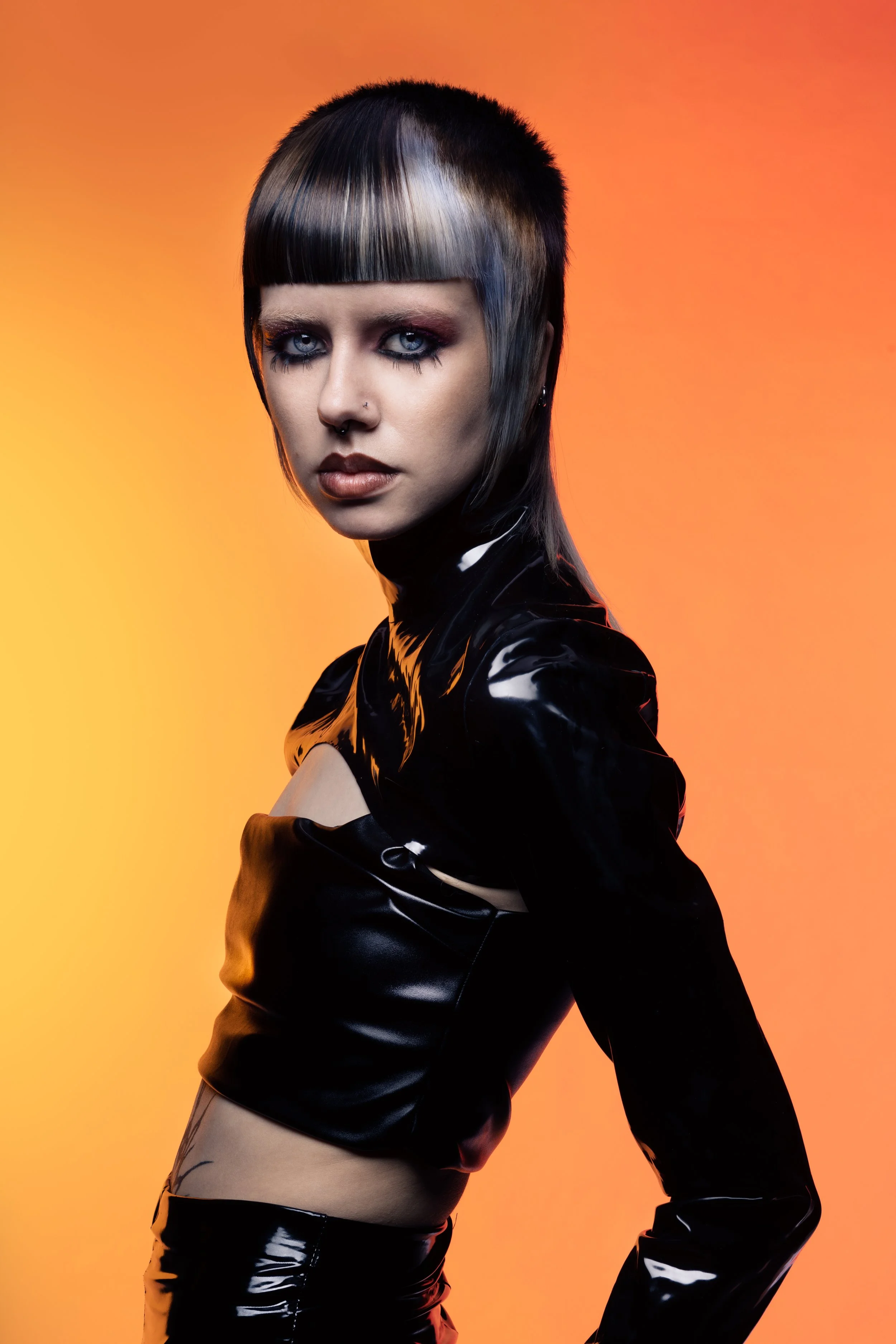 A woman with short, edgy black and gray hair, intense makeup, and piercings wearing a shiny black leather outfit, standing against a gradient orange background.