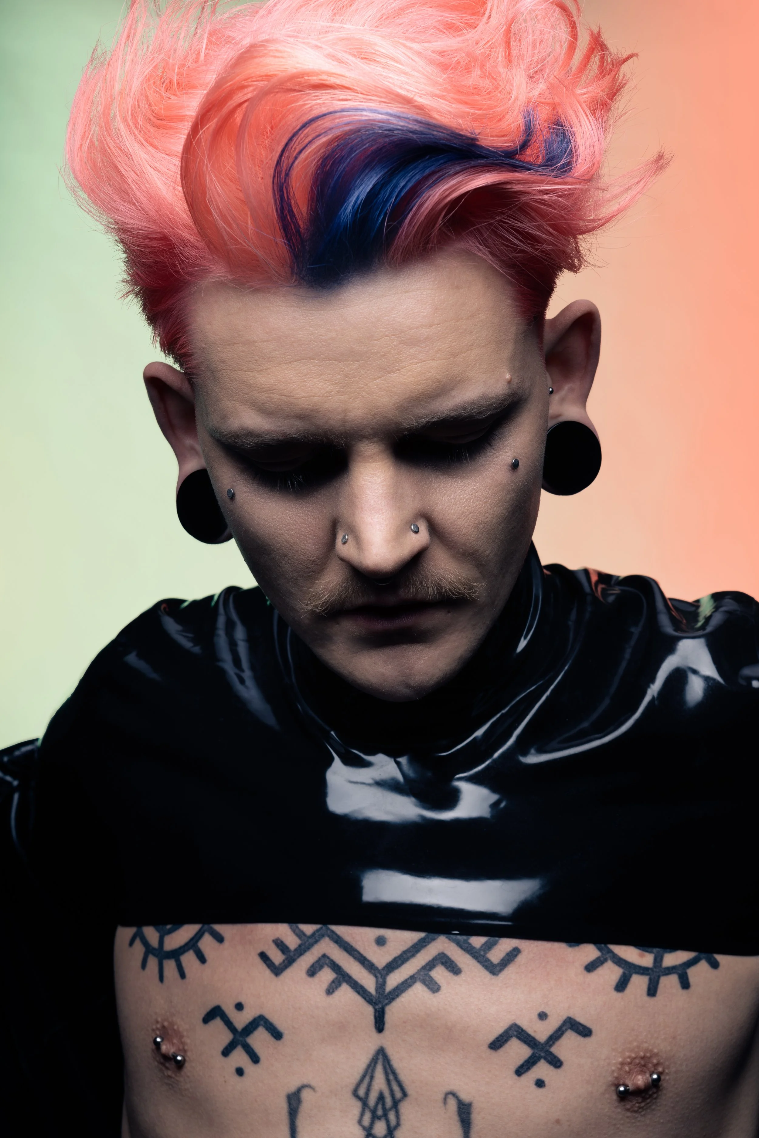 A person with colorful pink and blue hair styled upwards, wearing black clothing, with multiple facial piercings, large black earrings, and tattoos on their chest, looking downward against a gradient background.