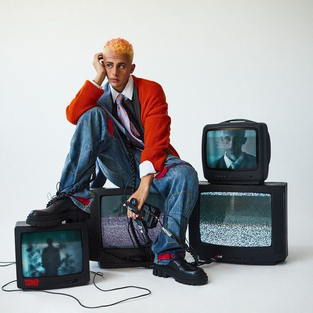 A young person with bleached blonde hair sitting on stacked vintage TVs, holding a video game controller, surrounded by screens displaying static or distorted images, in a minimalist setting.