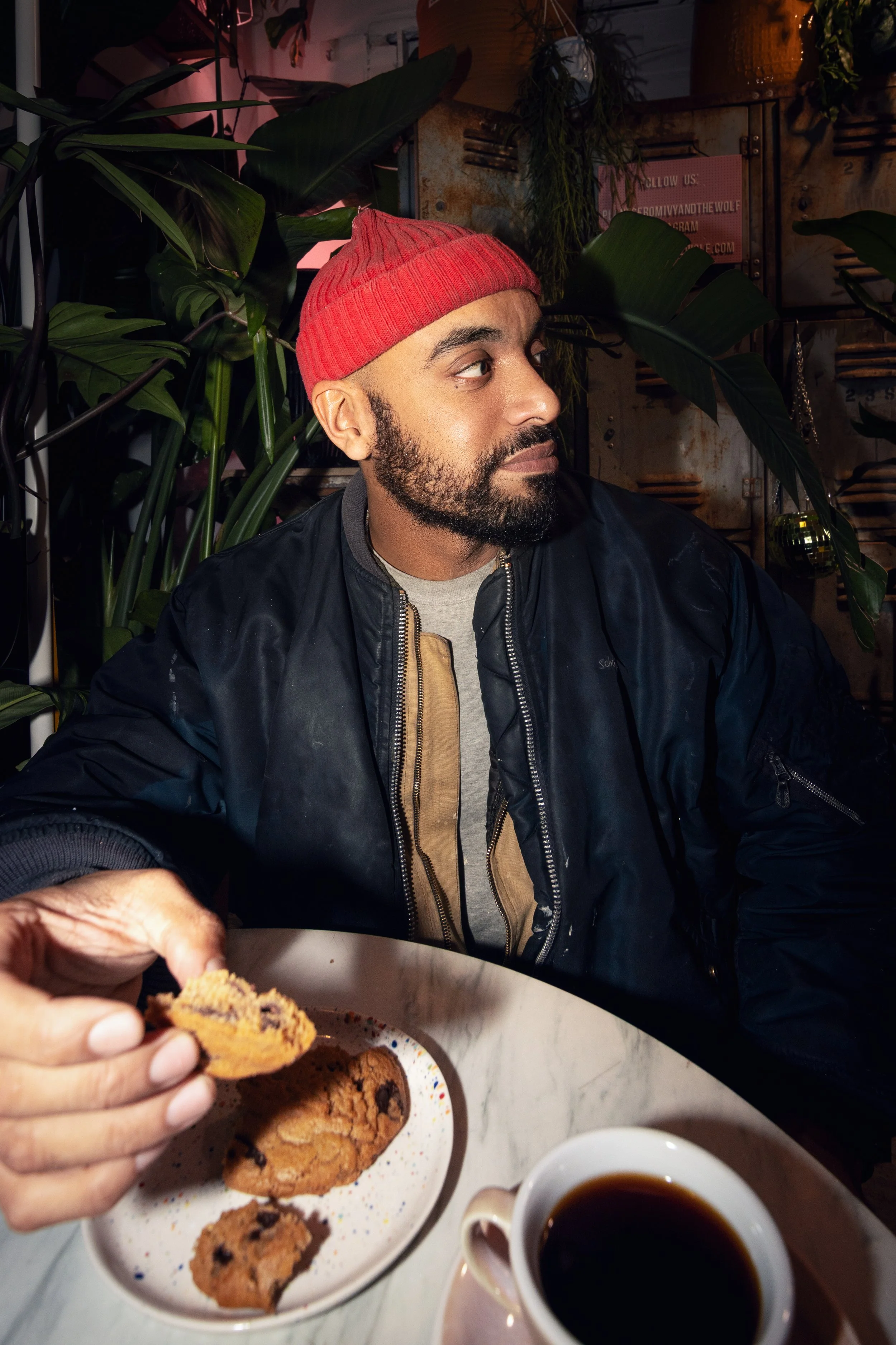 A man with a beard wearing a red knit beanie and a black jacket sits at a table with a cup of coffee and a plate of cookies. He is looking to his left, in a setting with green plants and metal lockers in the background.