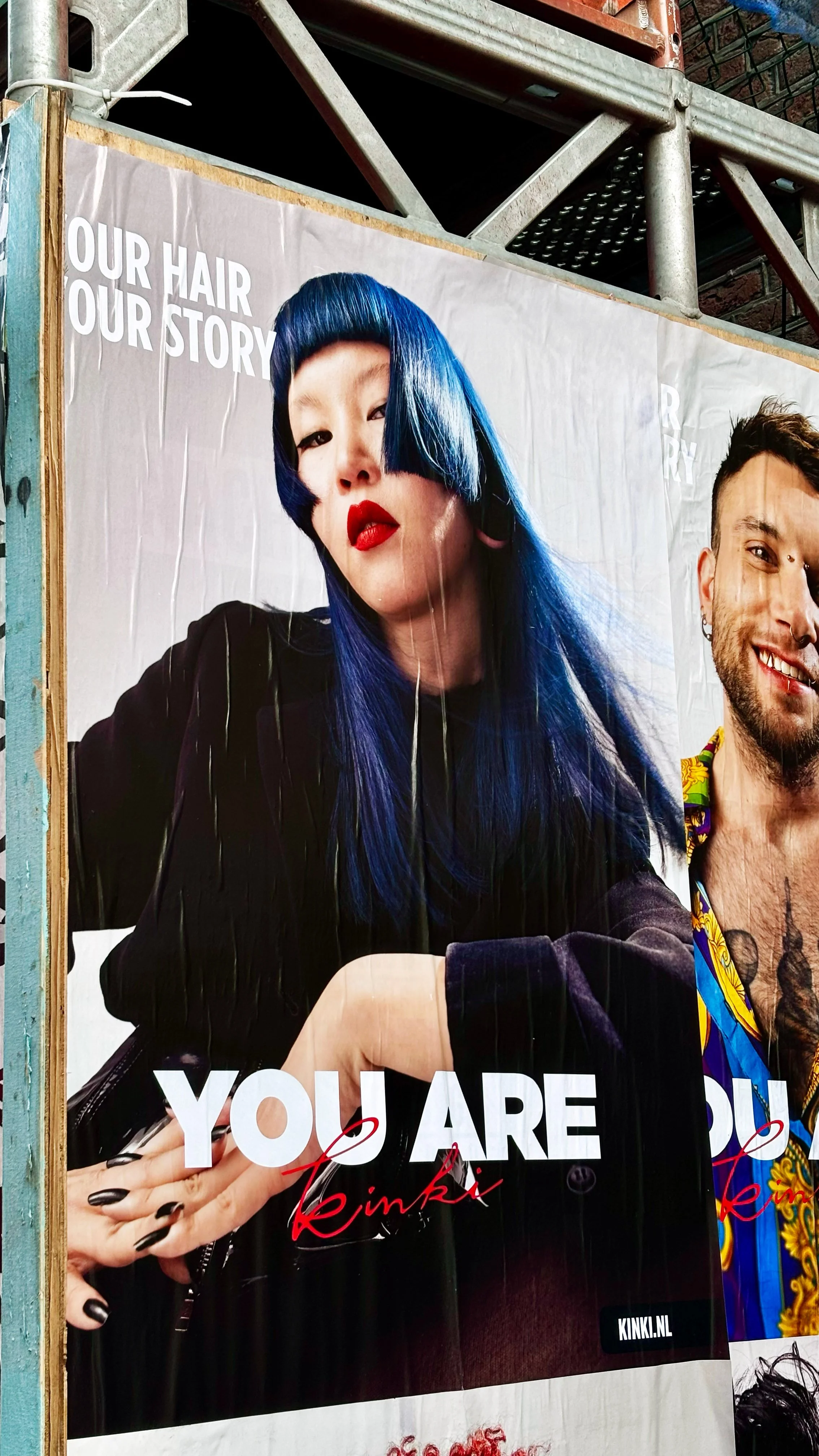 A woman with long blue hair, red lipstick, and black nails, wearing a black outfit, is looking confidently at the camera. The poster reads 'OUR HAIR OUR STORY YOU ARE YOU kinki'.