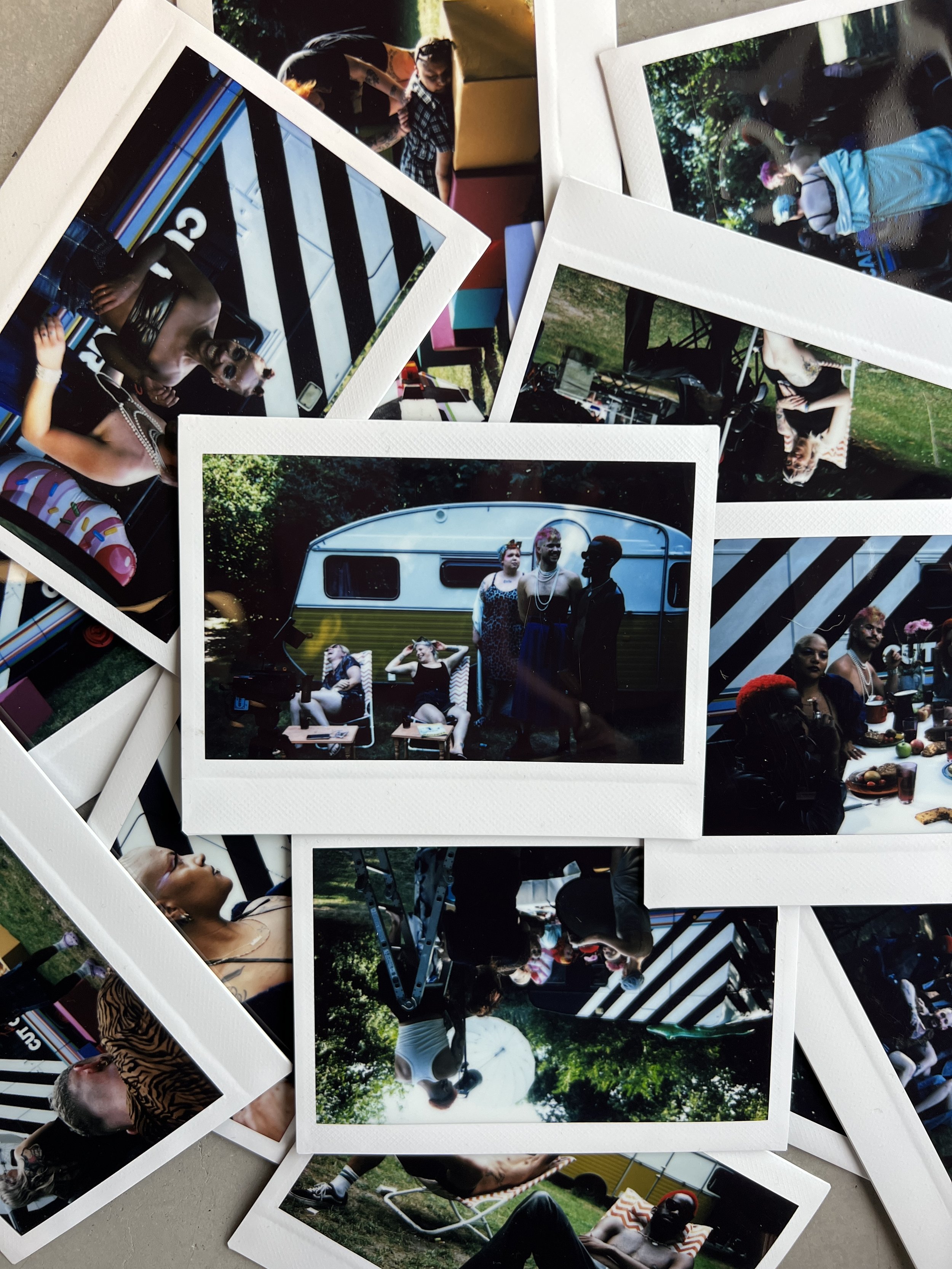A collection of Polaroid photos scattered on a surface, showcasing people at an outdoor gathering, including portraits, group shots, and candid moments with a vintage trailer in the background.