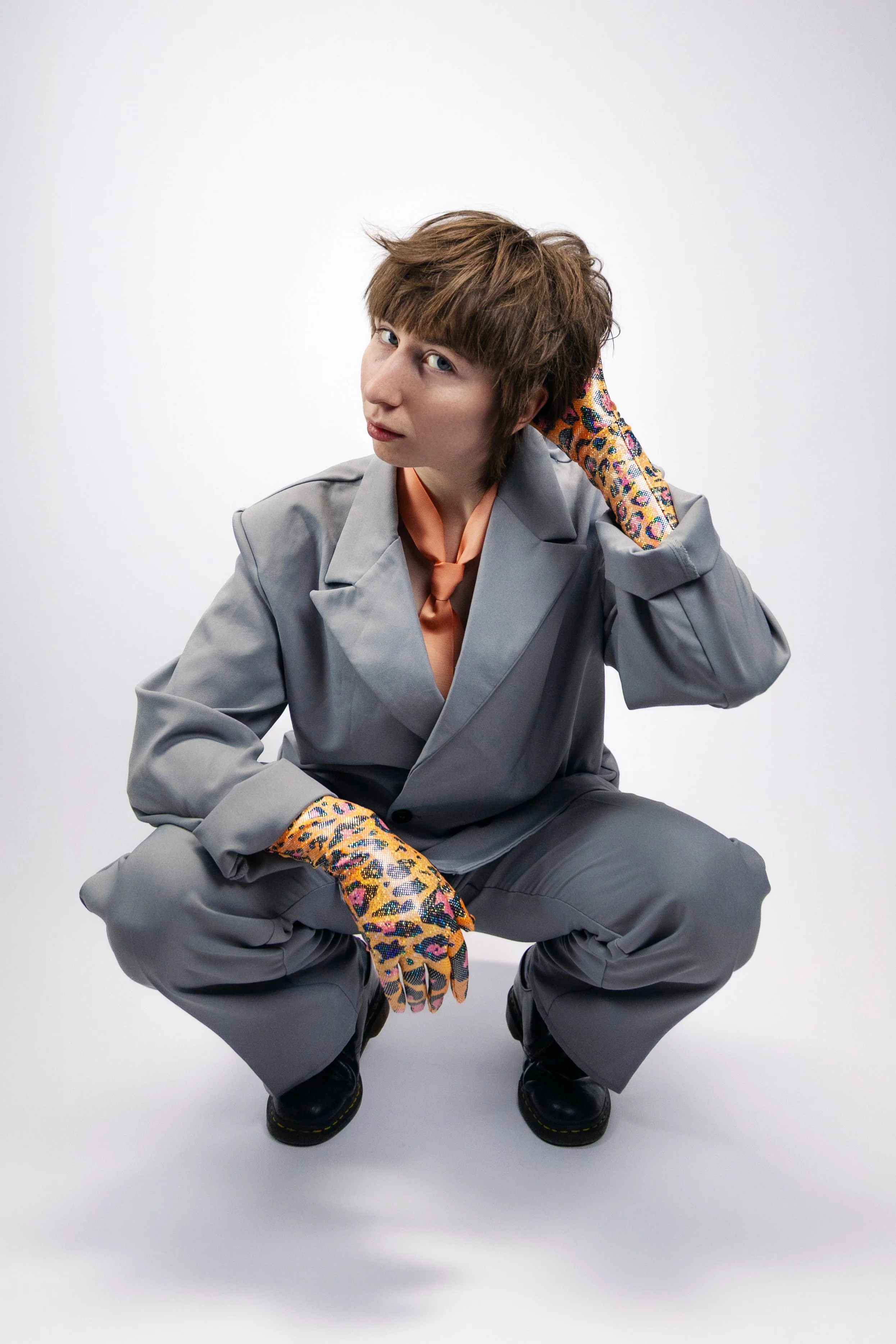 Person with short brown hair squatting, wearing a gray suit, colorful leopard print gloves, and black shoes, against a plain white background.