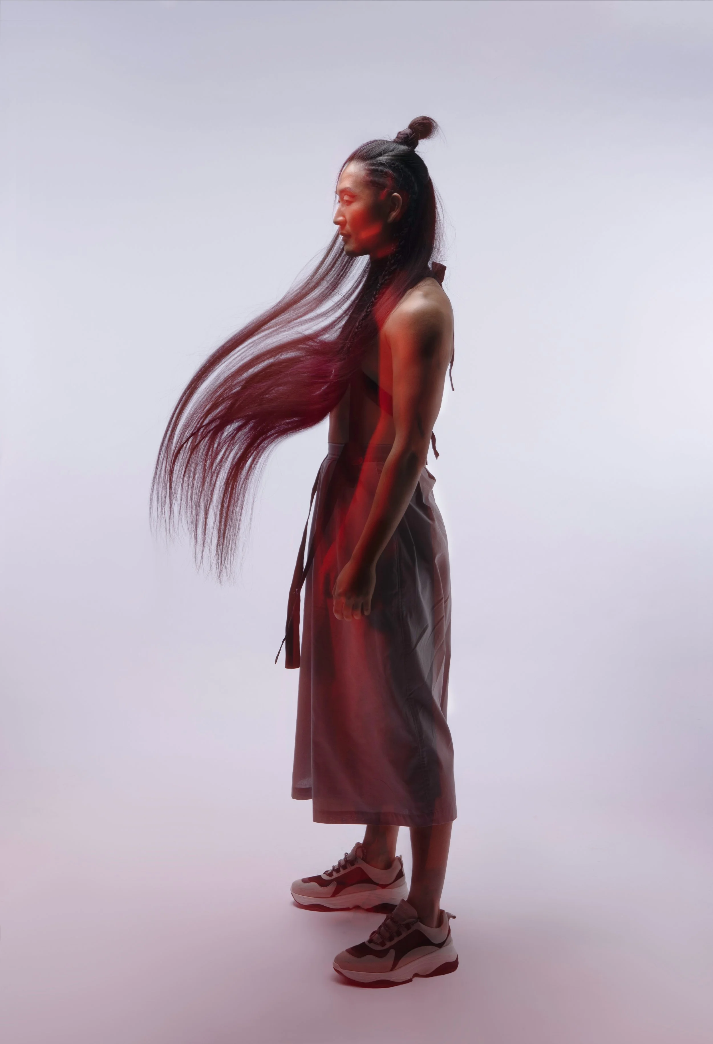 A woman with long, flowing hair standing sideways on a plain background, wearing athletic shoes and loose pants.