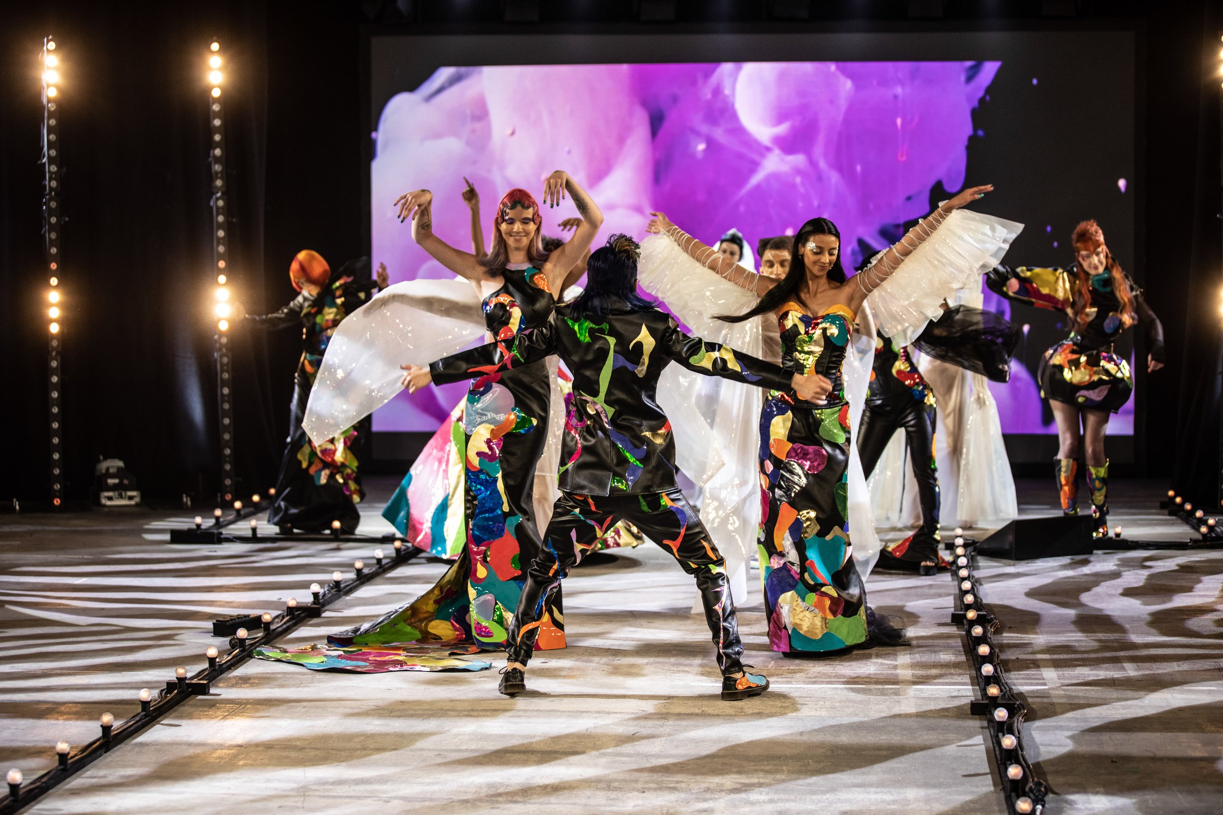Diverse group of performers on stage dancing in colorful, patterned costumes with large white wing-like accessories, with purple abstract background.
