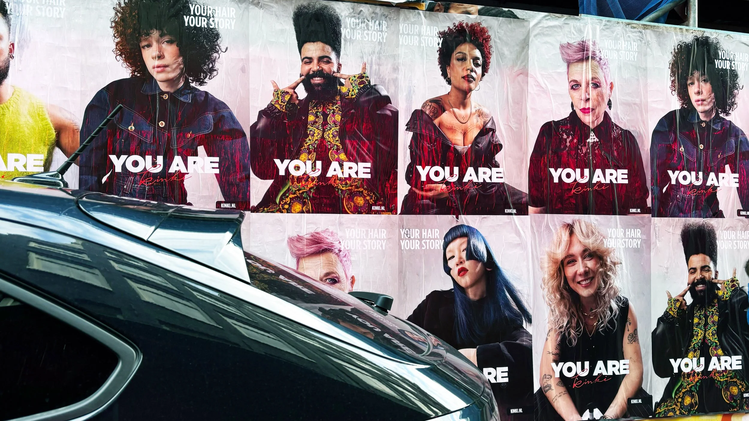 Colorful posters featuring diverse models with vibrant hairstyles and expressive poses, with the text "YOU ARE Rinki" and "YOUR HAIR YOUR STORY" in the background, along with a partially visible car in the foreground.