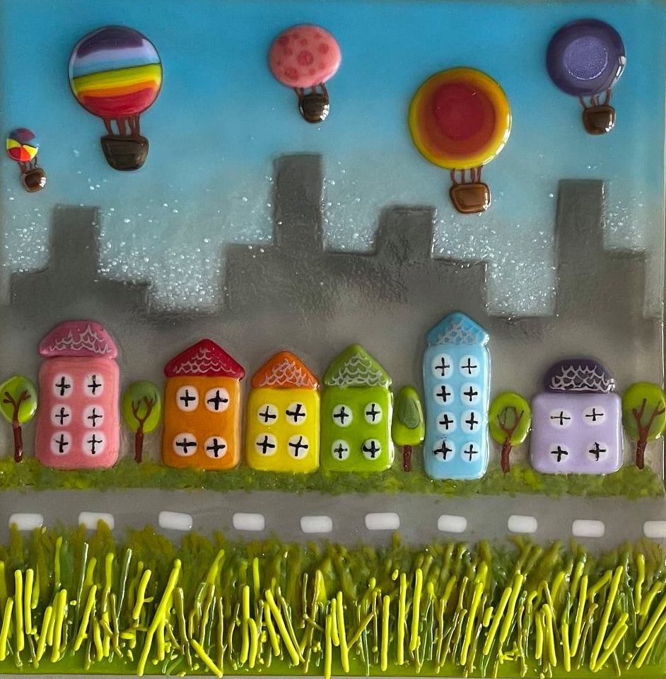 Colorful miniature houses with trees, hot air balloons in the sky, and a grassy foreground set against a city skyline background.
