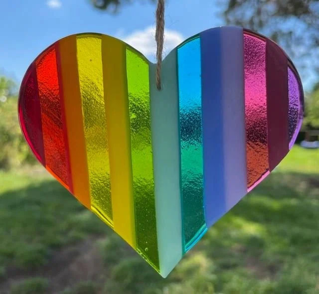 A colorful rainbow heart-shaped glass ornament hanging outdoors with a garden and trees in the background.