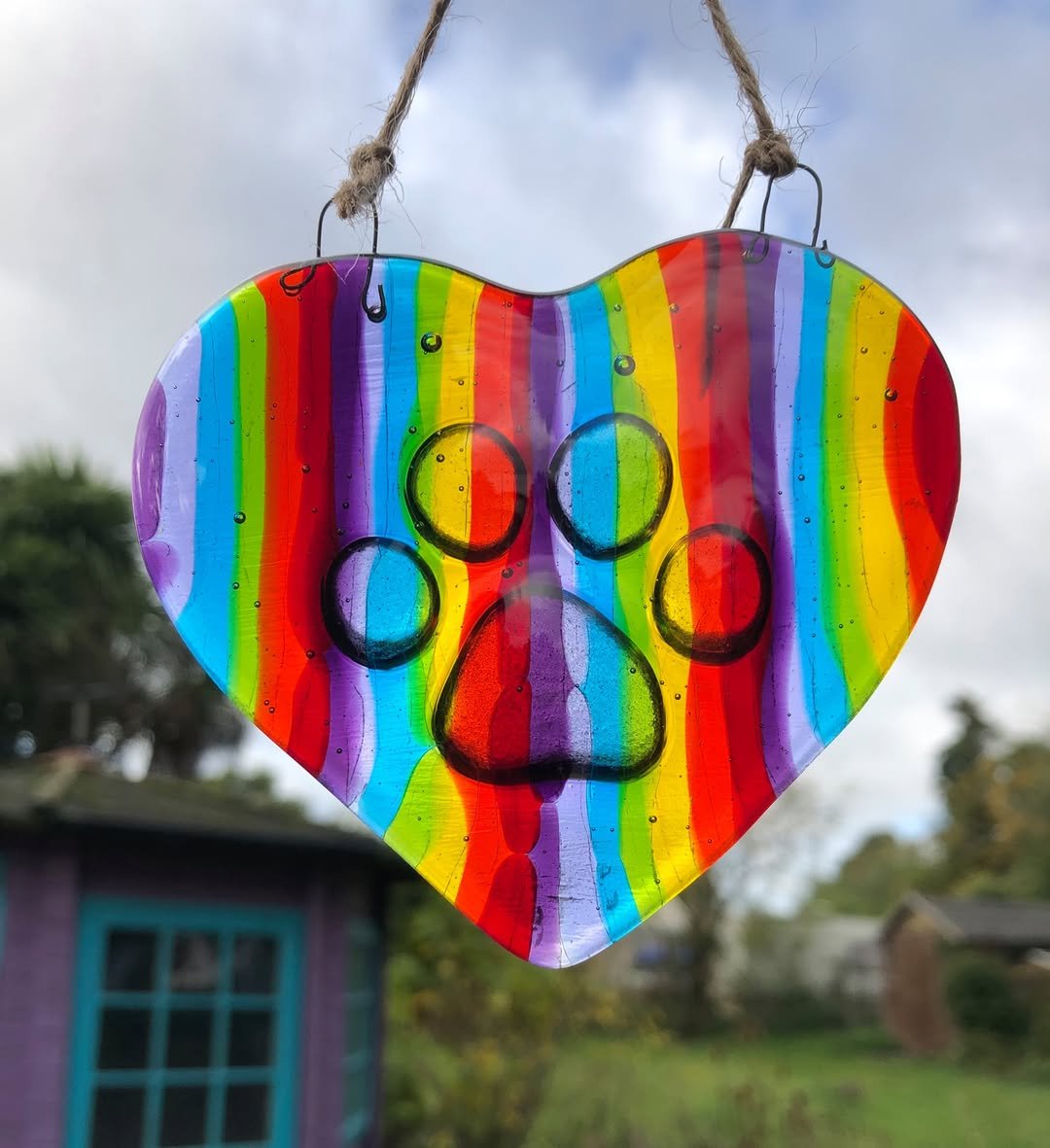 A colorful heart-shaped ornament with rainbow stripes, hanging outdoors with a cloudy sky in the background.