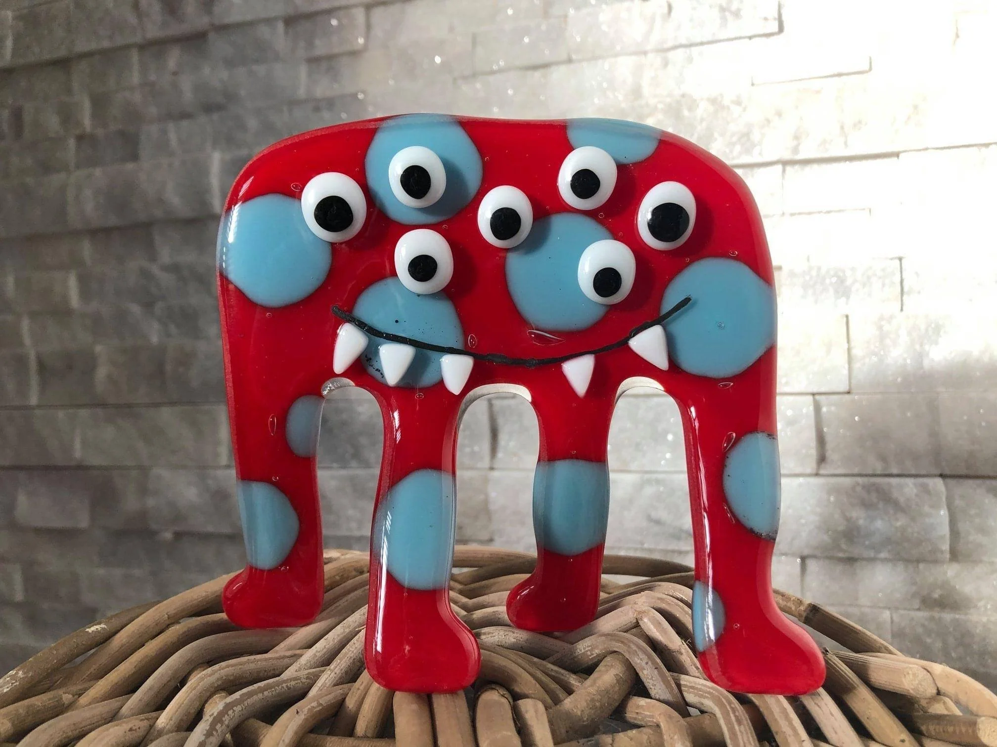 Decorative glass elephant with a red body, blue spots, and multiple googly eyes, sitting on a woven surface with a gray stone wall background.
