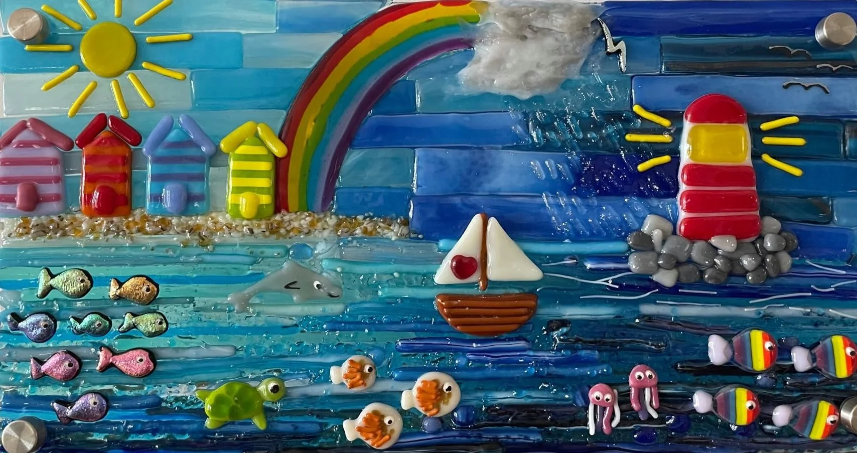 Colorful mosaic art depicting a seaside scene with a yellow sun, rainbow, houses, dolphins, fish, a sailboat, a lighthouse, and other ocean elements using glass and stone materials.
