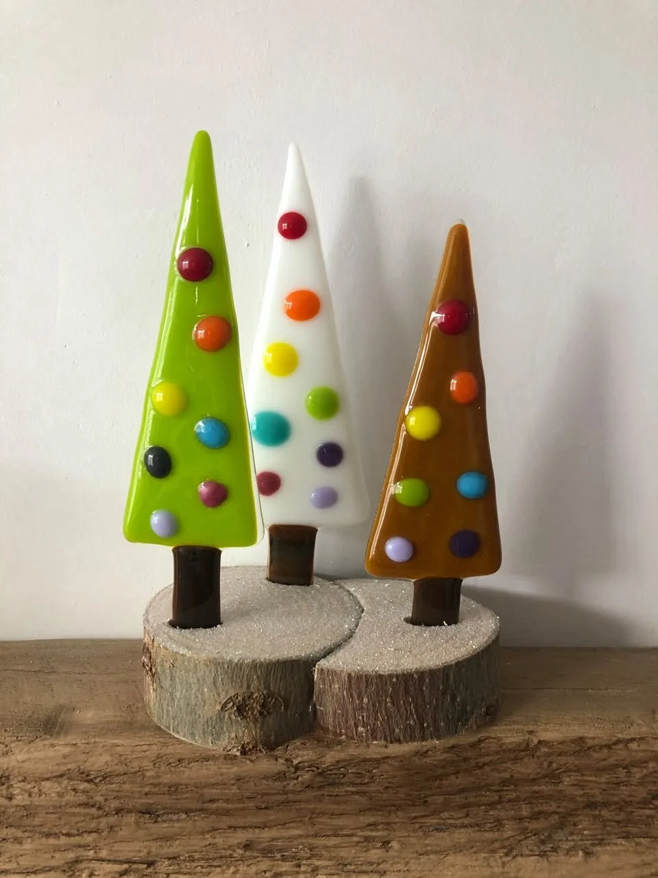 Colorful glass Christmas trees on wooden bases, placed on a wooden surface against a plain background.