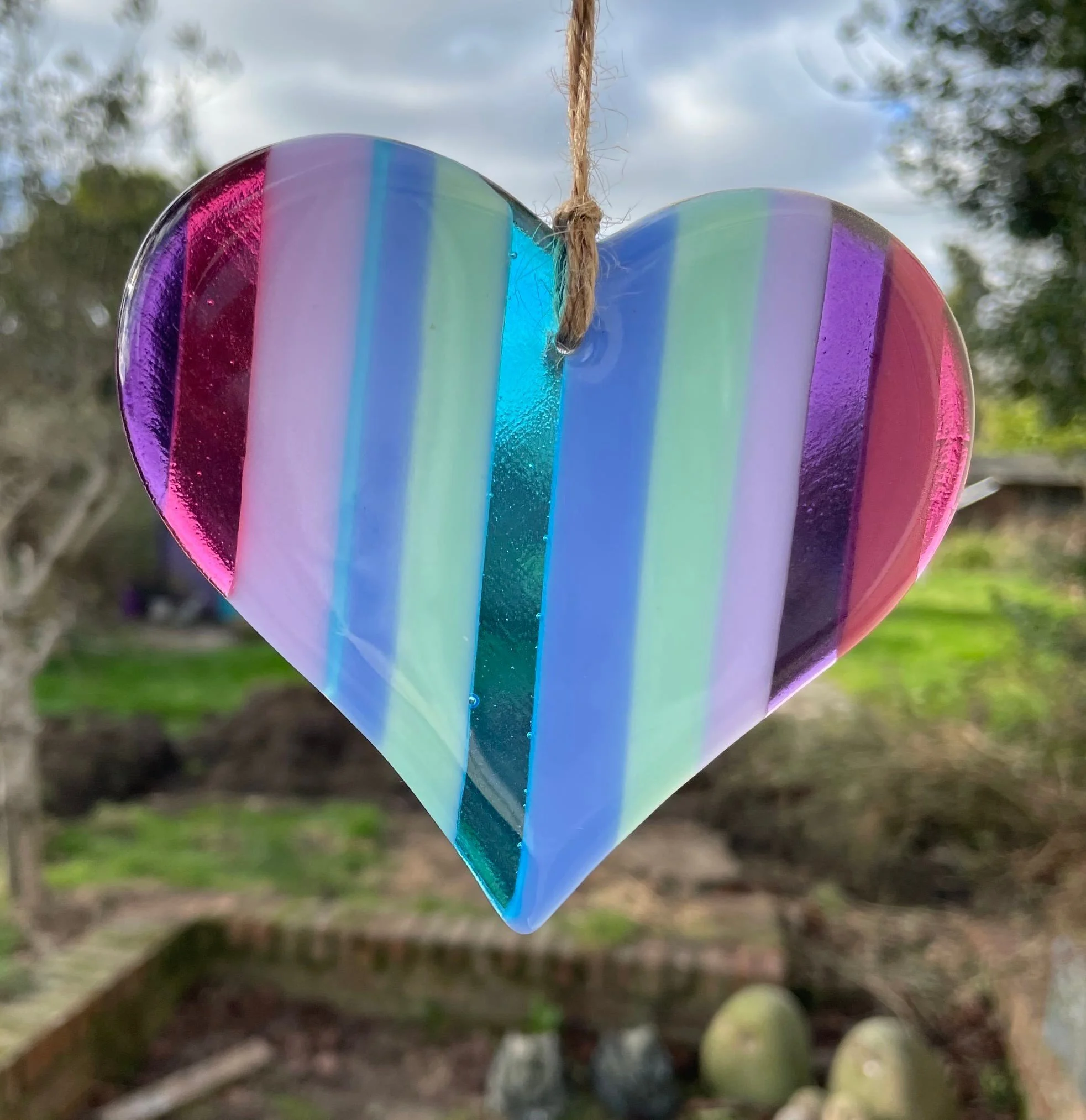 Colorful glass heart hanging outdoors with a blurred background of trees and sky.