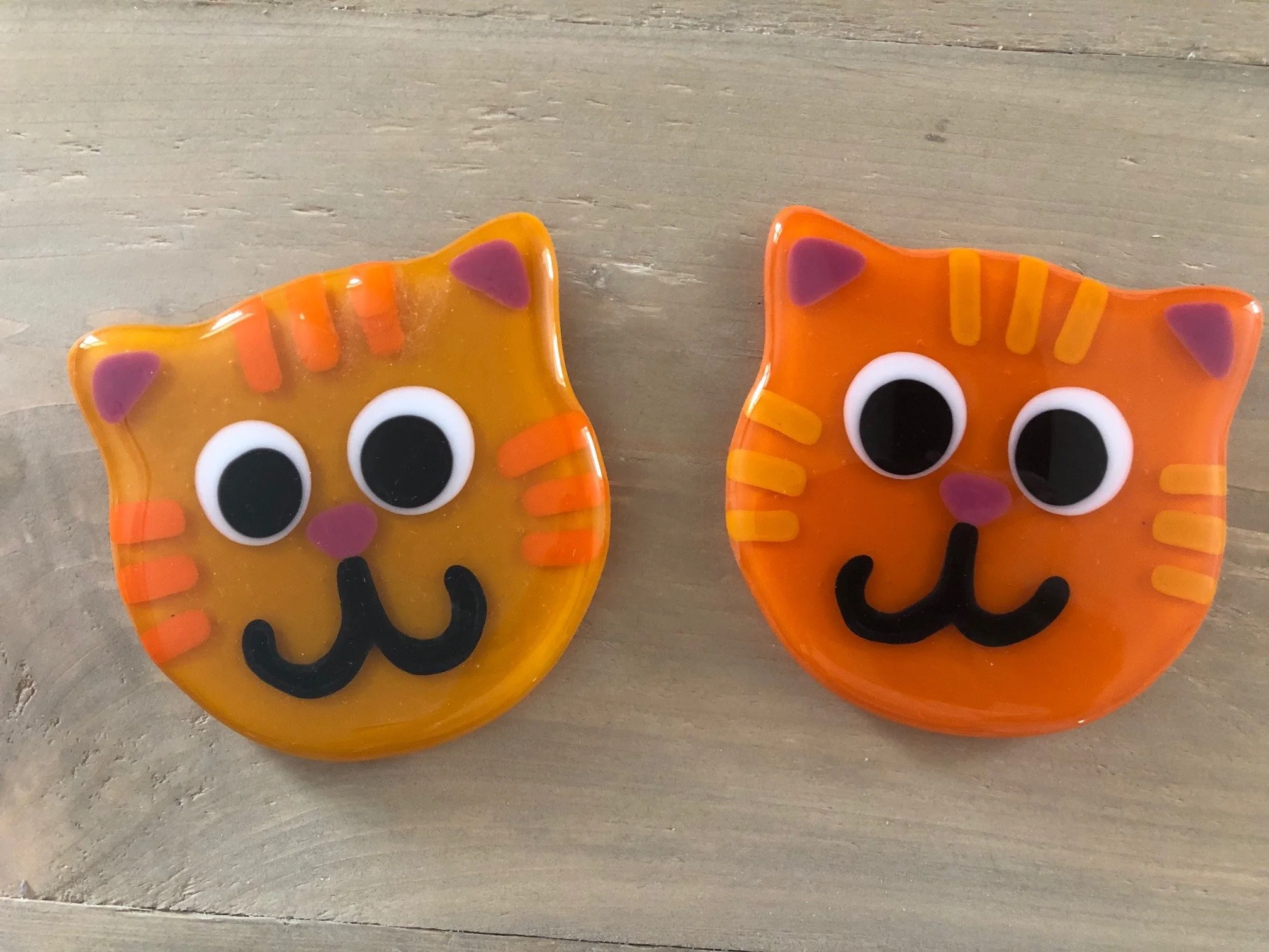 Two colorful ceramic cat-shaped dishes with large googly eyes, purple ears, and black smiling mouths, placed on a wooden surface.