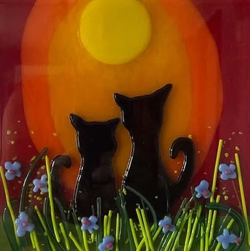 Glass art of two black cats sitting on grass under a yellow sun with an orange background.