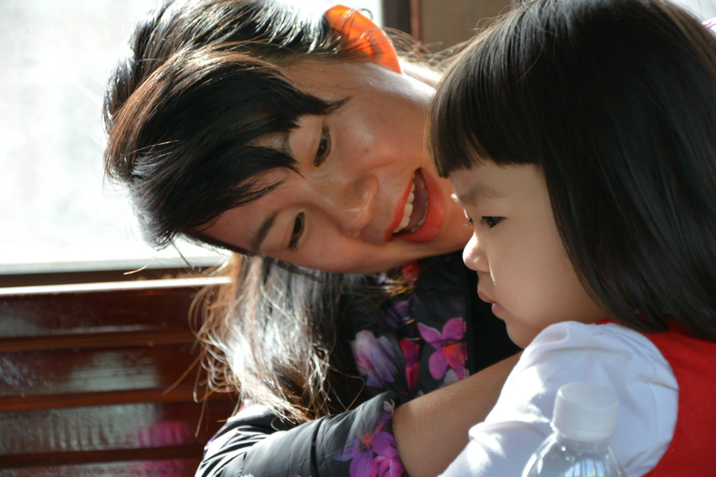 An adult woman with dark hair and red lipstick leaning close to a young girl with dark hair and bangs, sitting at a table near a window, with a water bottle in front of the girl.