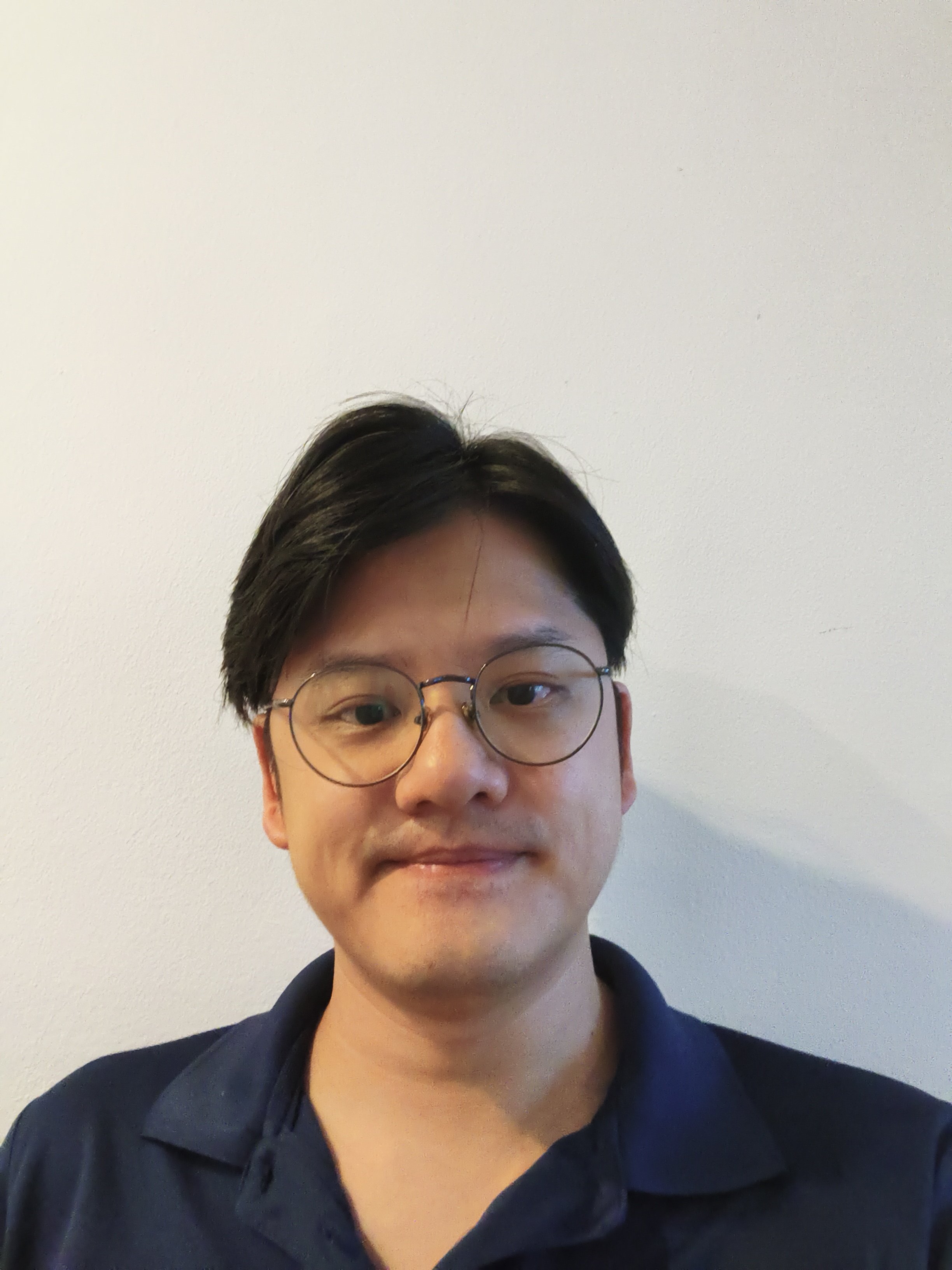 A young man with black hair, glasses, and a navy blue shirt standing against a plain white wall.
