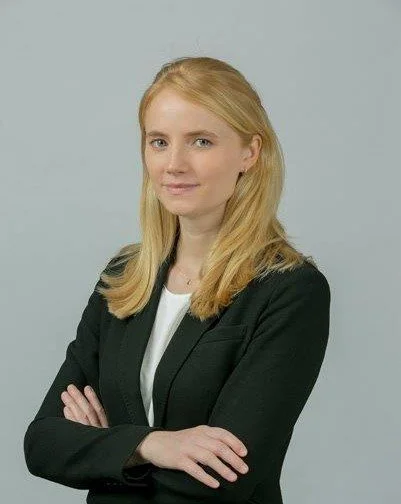 A woman with blonde hair wearing a black blazer and white top standing against a light gray background.