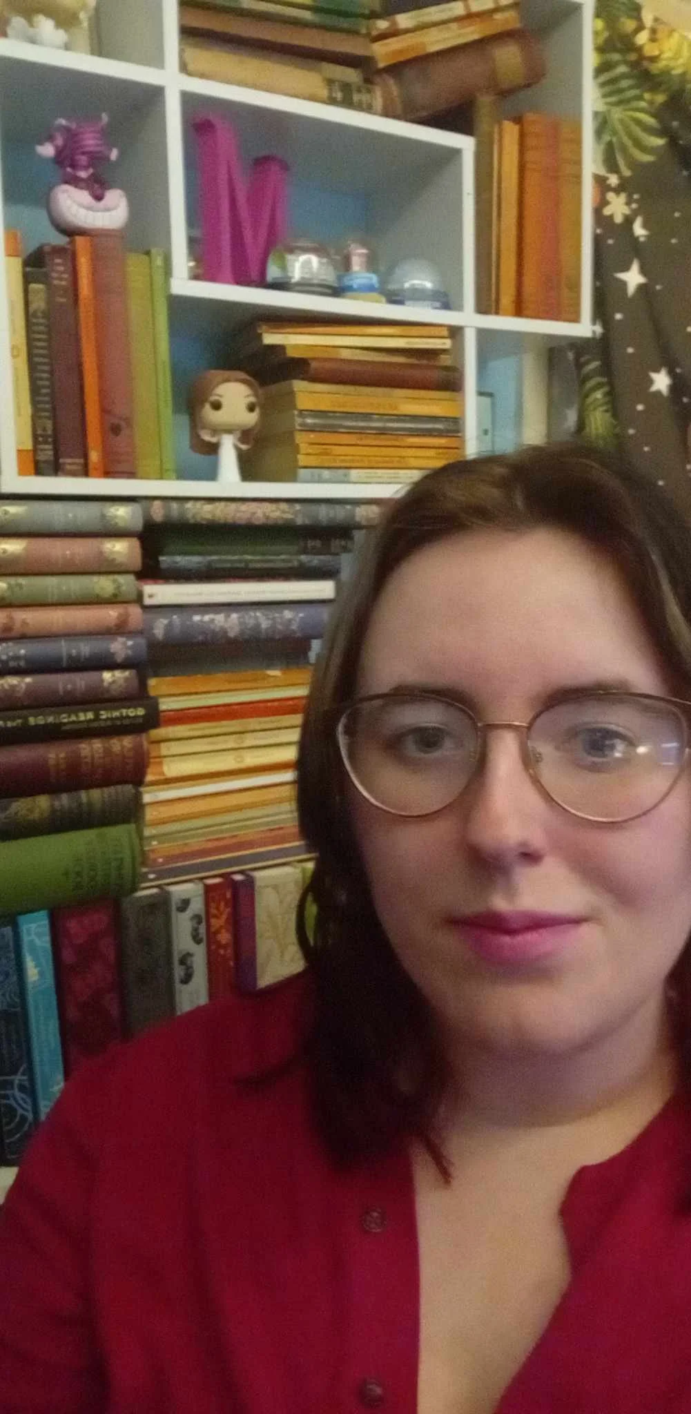 A woman with glasses and dark hair taking a selfie in front of a bookshelf filled with colorful books and decorative items.