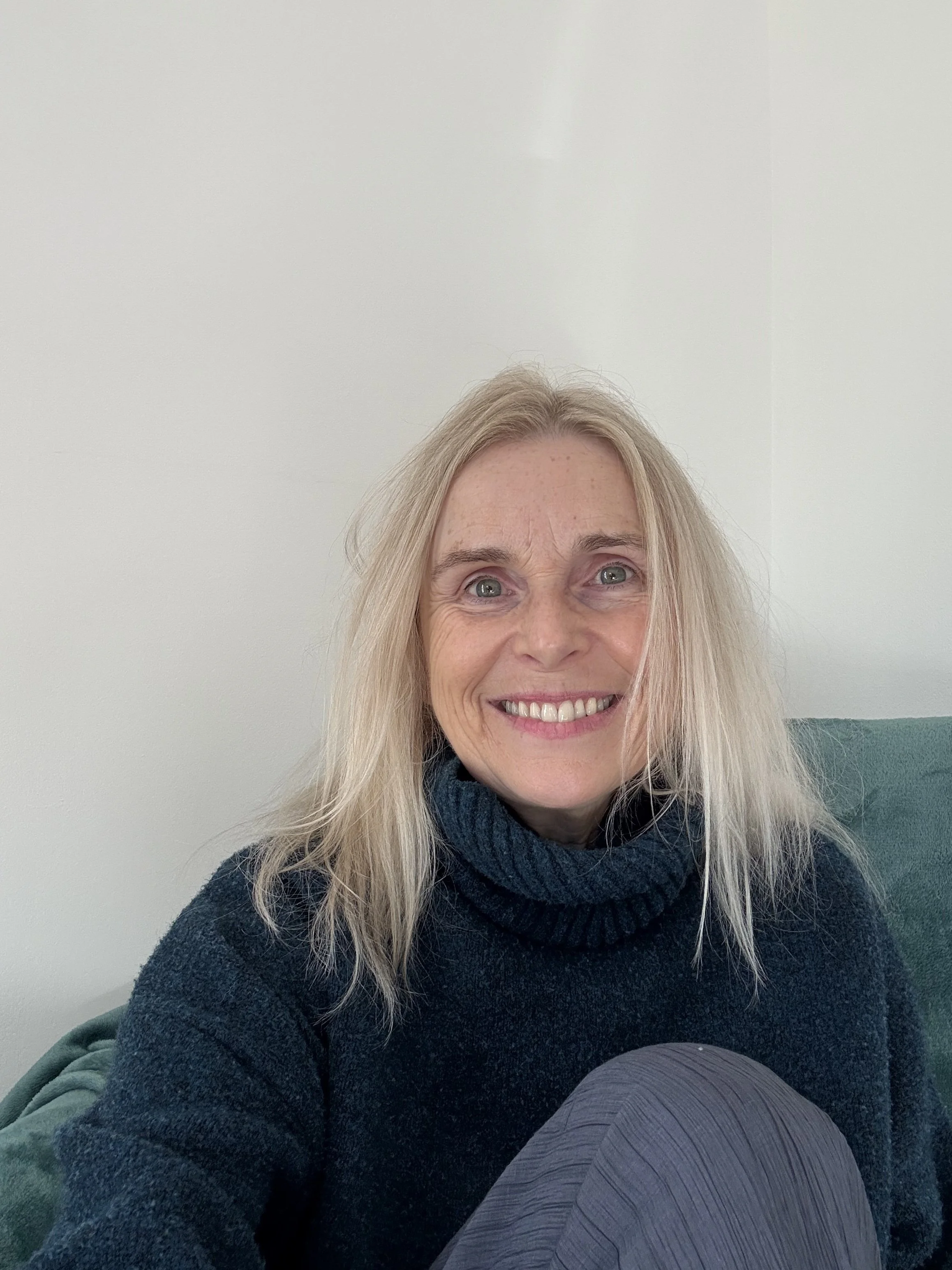 A woman with blonde hair, blue eyes, and a wide smile, wearing a dark turtleneck sweater, sitting on a green velvet chair against a plain white wall.