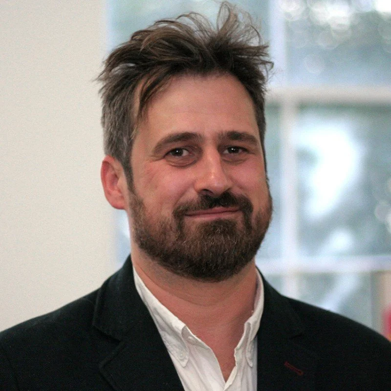 A man with a beard and tousled hair wearing a white shirt and black blazer, smiling softly, standing indoors with a window in the background.