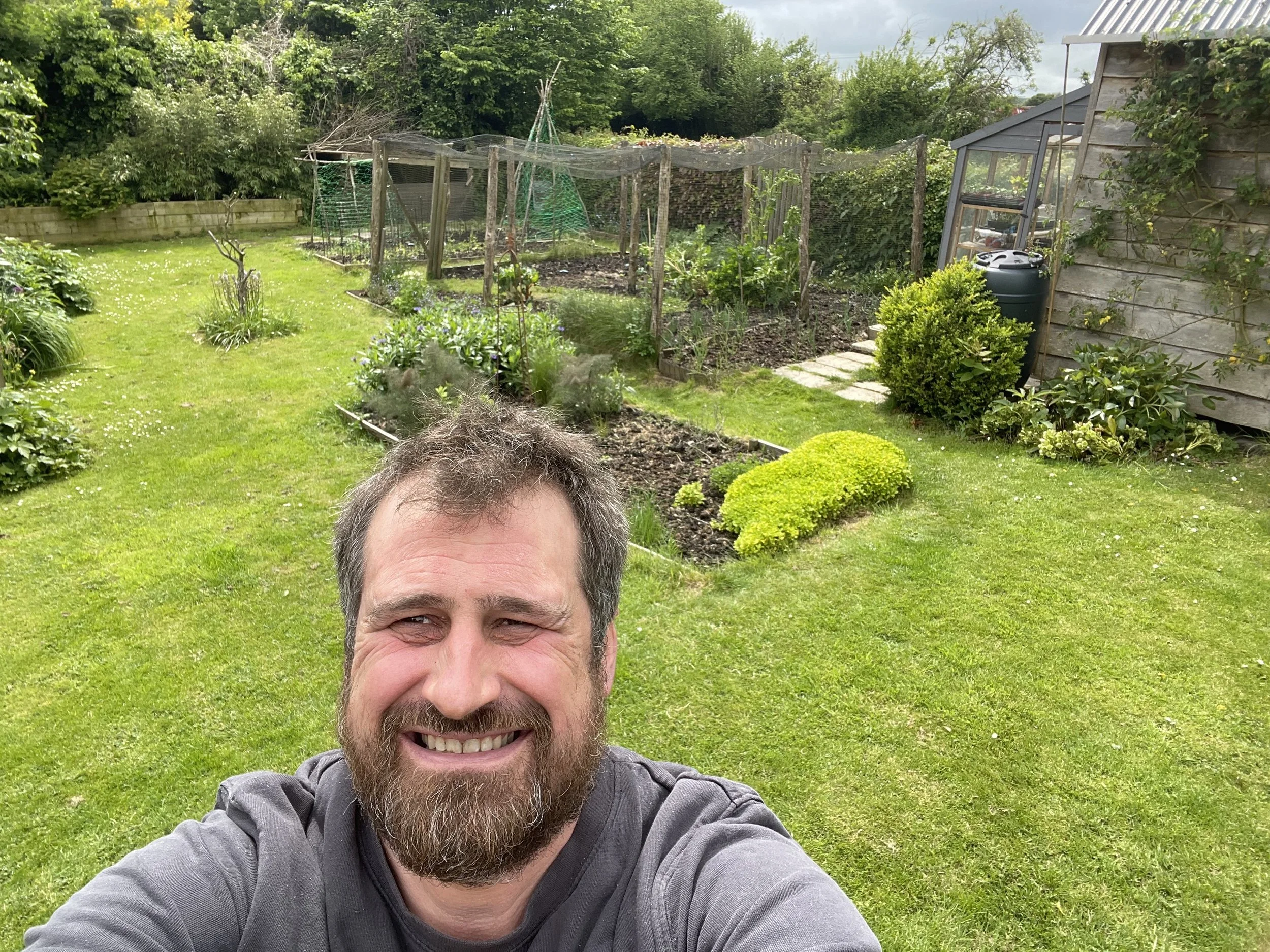 A man smiling and taking a selfie in a backyard garden with lush green grass, flower beds, a small shed, and a greenhouse in the background.