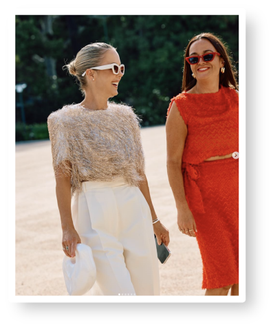 Two women wearing stylish outfits, sunglasses, and smiling, standing outdoors on a sunny day.