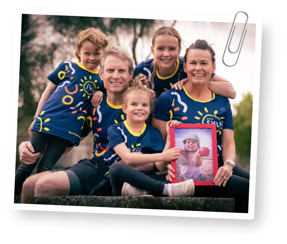Family of five wearing matching blue t-shirts with colorful designs, holding a framed photo of a girl in a bathing suit at the beach, outdoors in a park-like setting.
