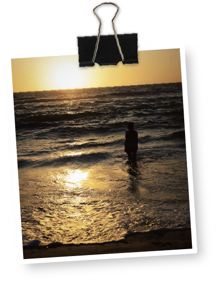 A person standing in the ocean at sunset, with waves and the sun low on the horizon, hanging from a black binder clip