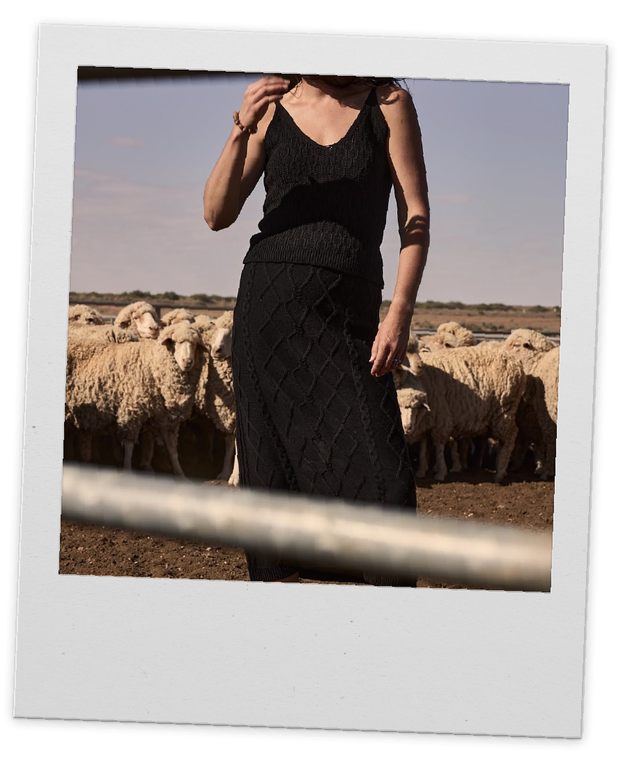 A woman wearing a black knitted sleeveless top and matching skirt standing in front of a flock of sheep on a farm during daytime.