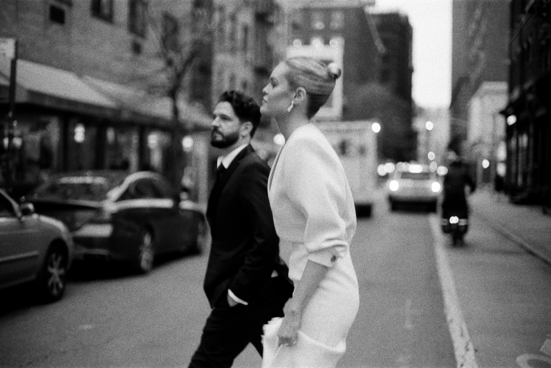 A black and white photo of a man and a woman walking on the street in a city. The woman is wearing a white blazer and has her hair in an updo, while the man is in a dark suit with a beard. In the background, there are parked cars and blurred buildings, with some pedestrians and a scooter on the street.