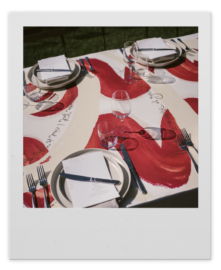 A table set for a meal with plates, silverware, napkins, and glasses on an outdoor tablecloth with red and cream abstract design.