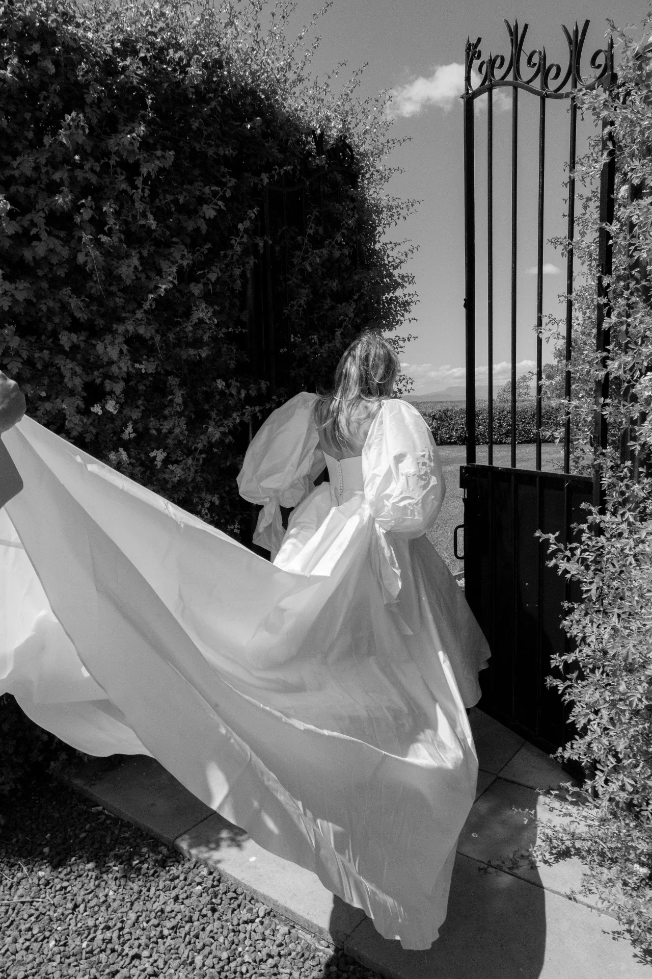 A woman wearing a wedding dress is walking through an open gate, with her dress flowing behind her. There is a large bush on the left and an open landscape in the background.