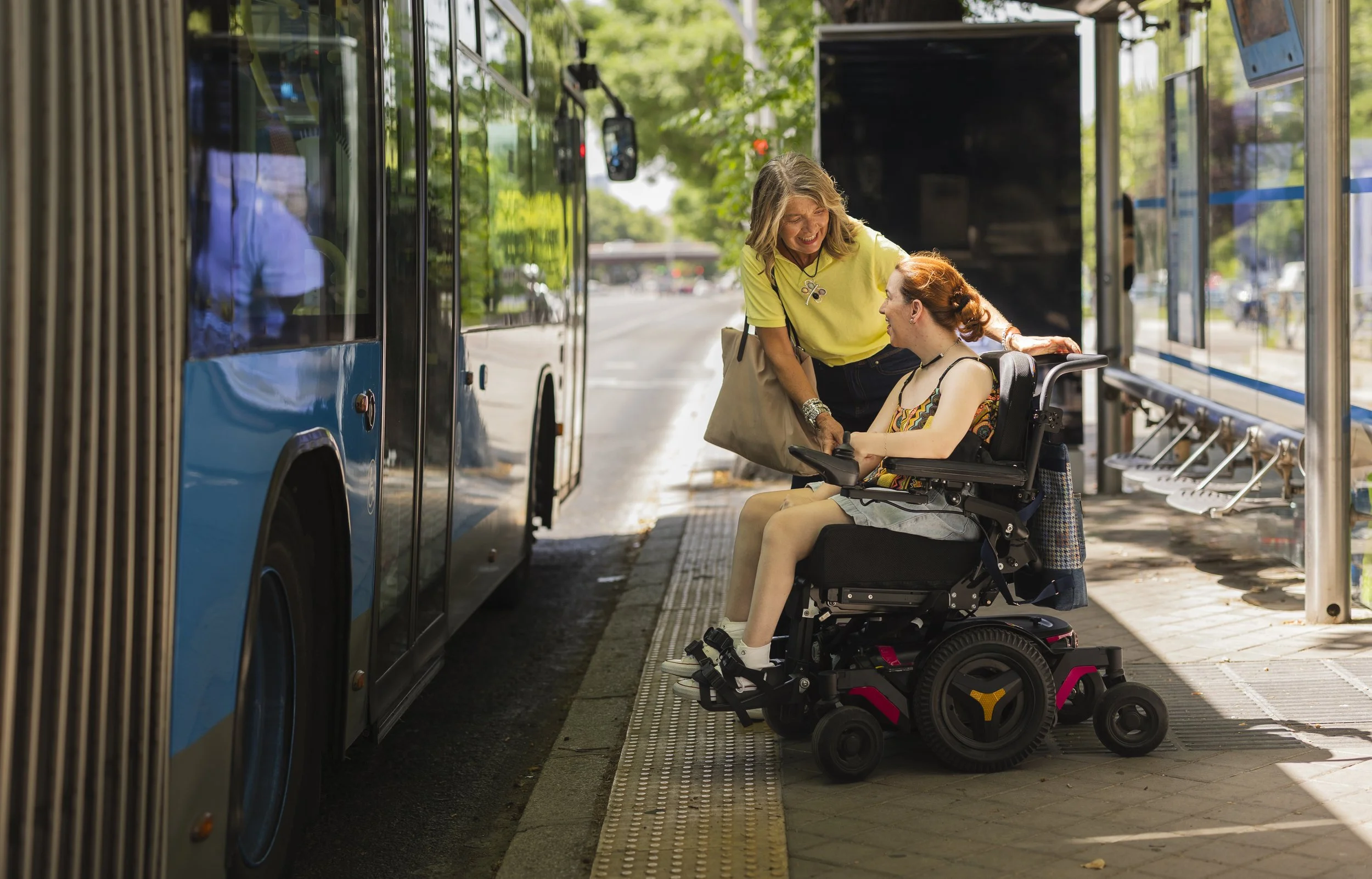 A woman in a wheelchair and an older woman are smiling and talking at a bus stop on a sunny day.