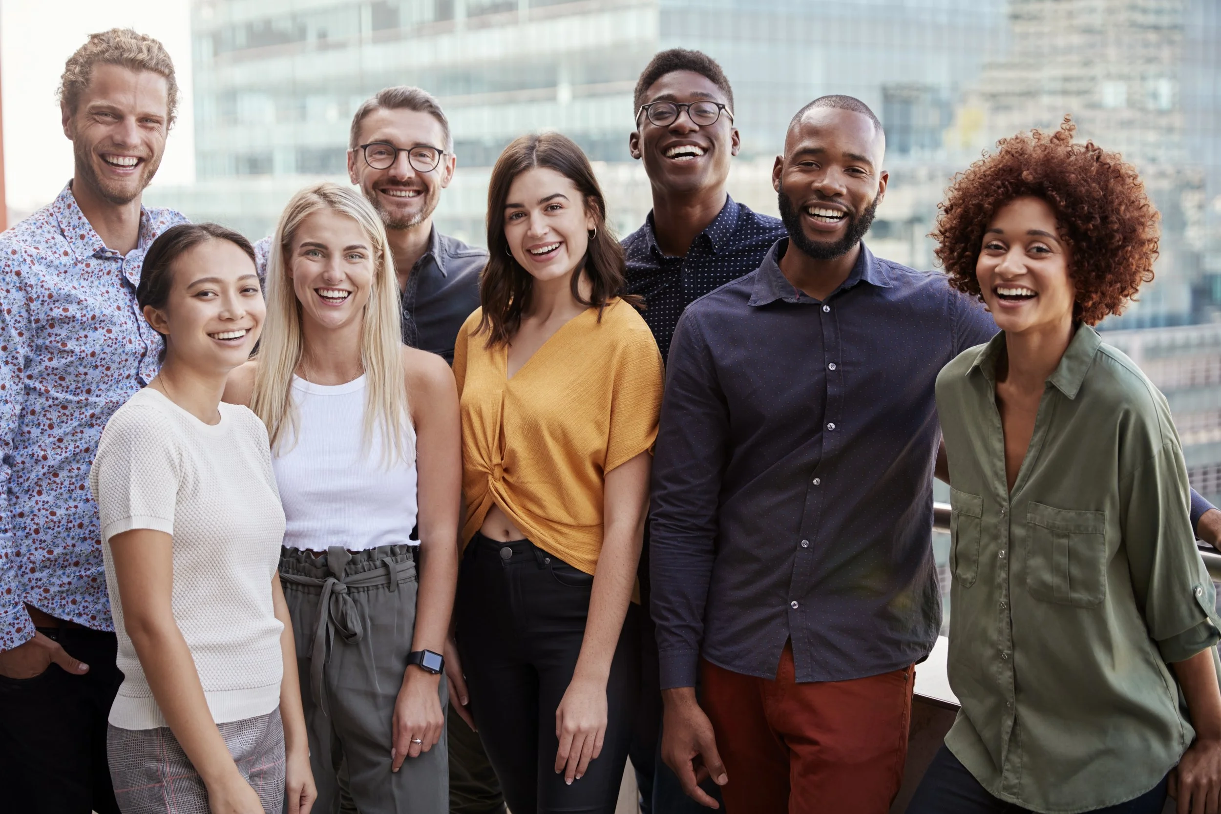 A group of nine diverse young adults smiling and posing outdoors in front of a cityscape with tall glass buildings.