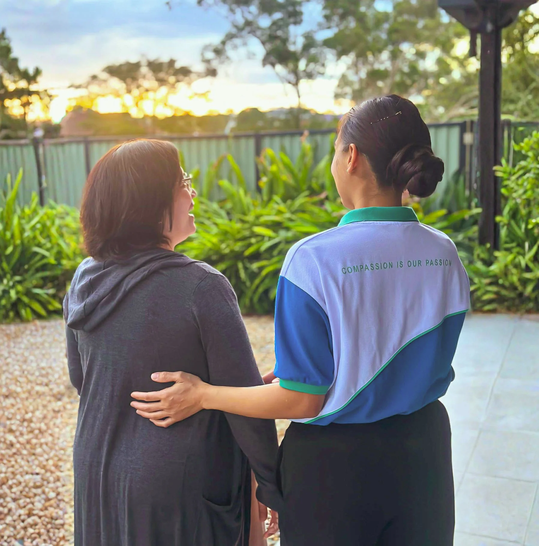 Two women standing outdoors in a garden at sunset, with one wearing a black hoodie and glasses, and the other in a white and blue uniform with the slogan "Compassion is our passion" on the back, engaging in a friendly conversation.