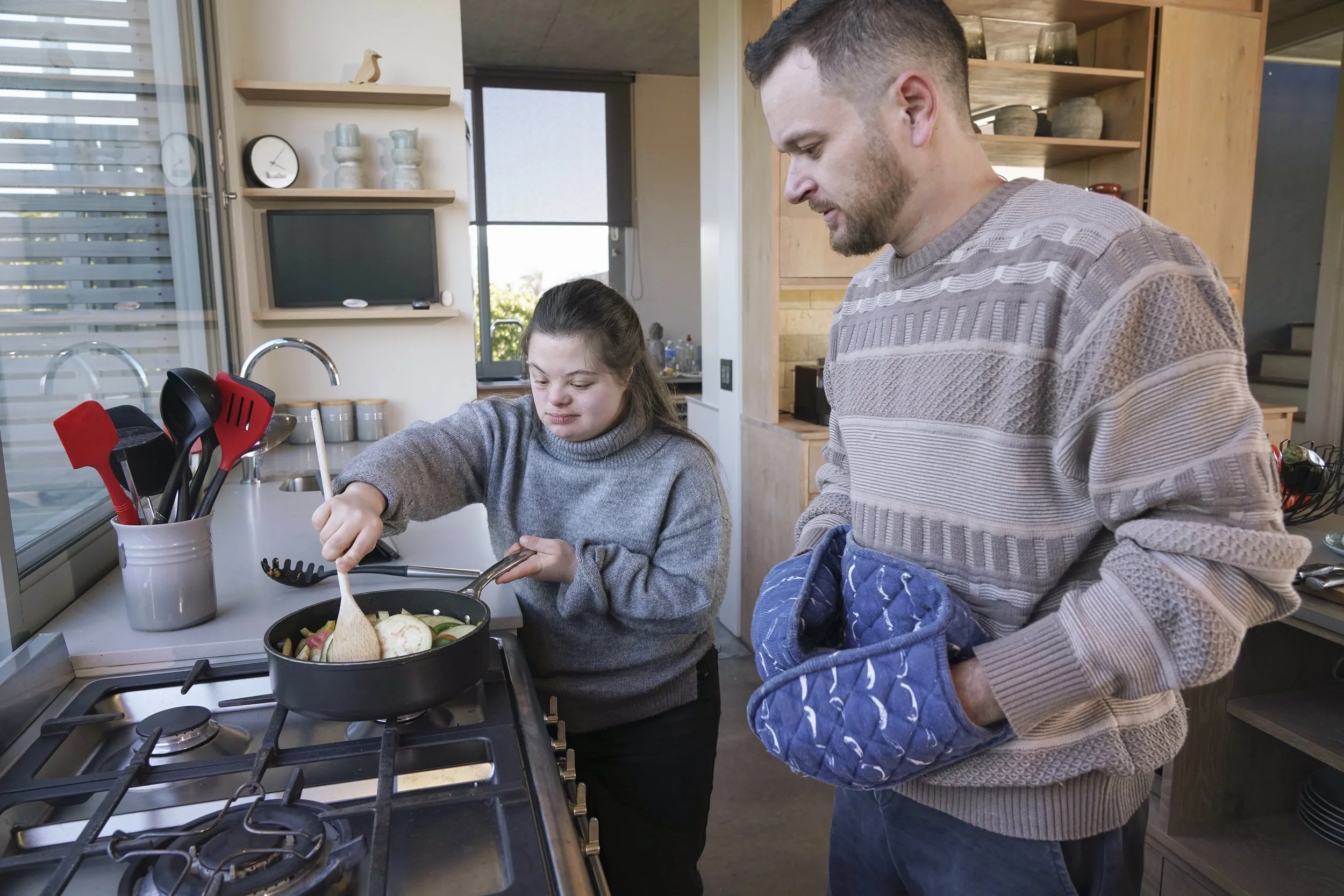 A man and girl cooking breakfast in a modern kitchen, girl stirring a frying pan with vegetables, man watching, wearing an oven mitts.