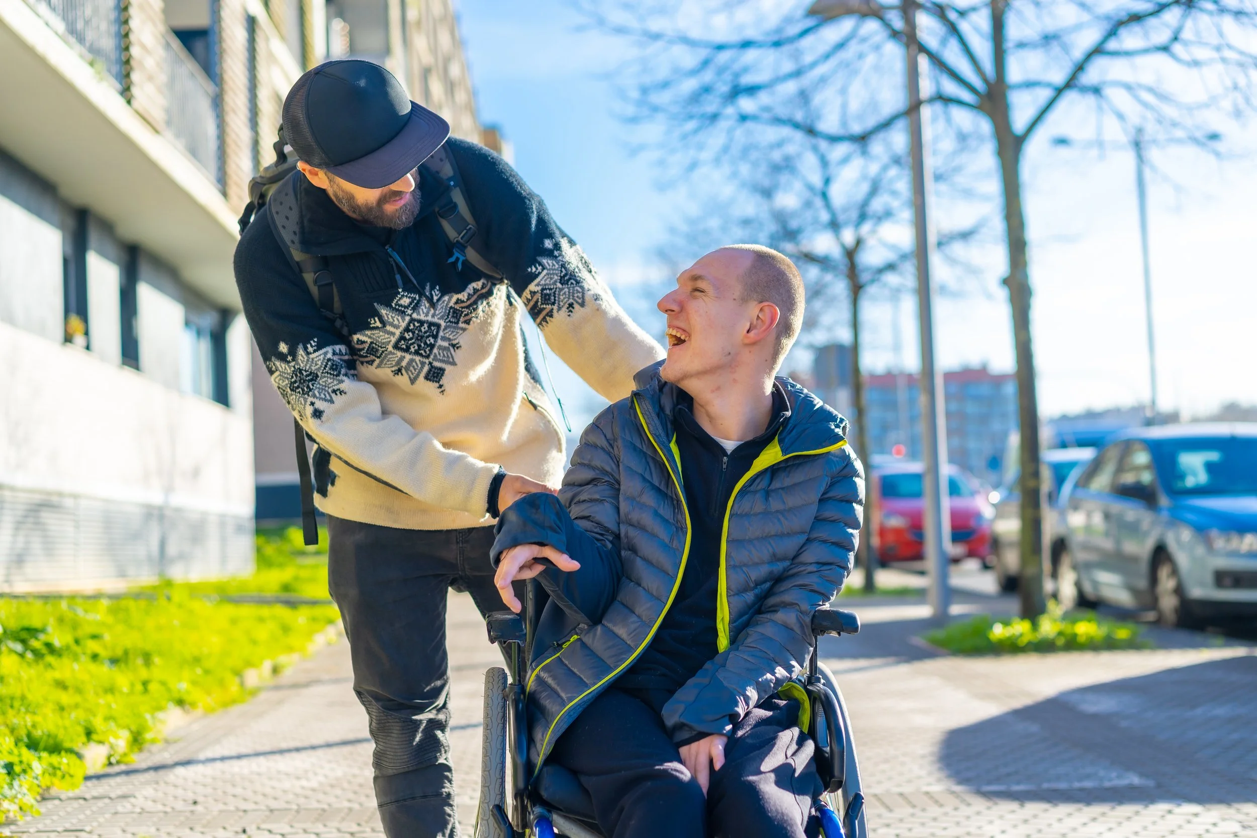 A man in a wheelchair smiling at a man standing next to him outdoors on a sunny day, with parked cars and buildings in the background.